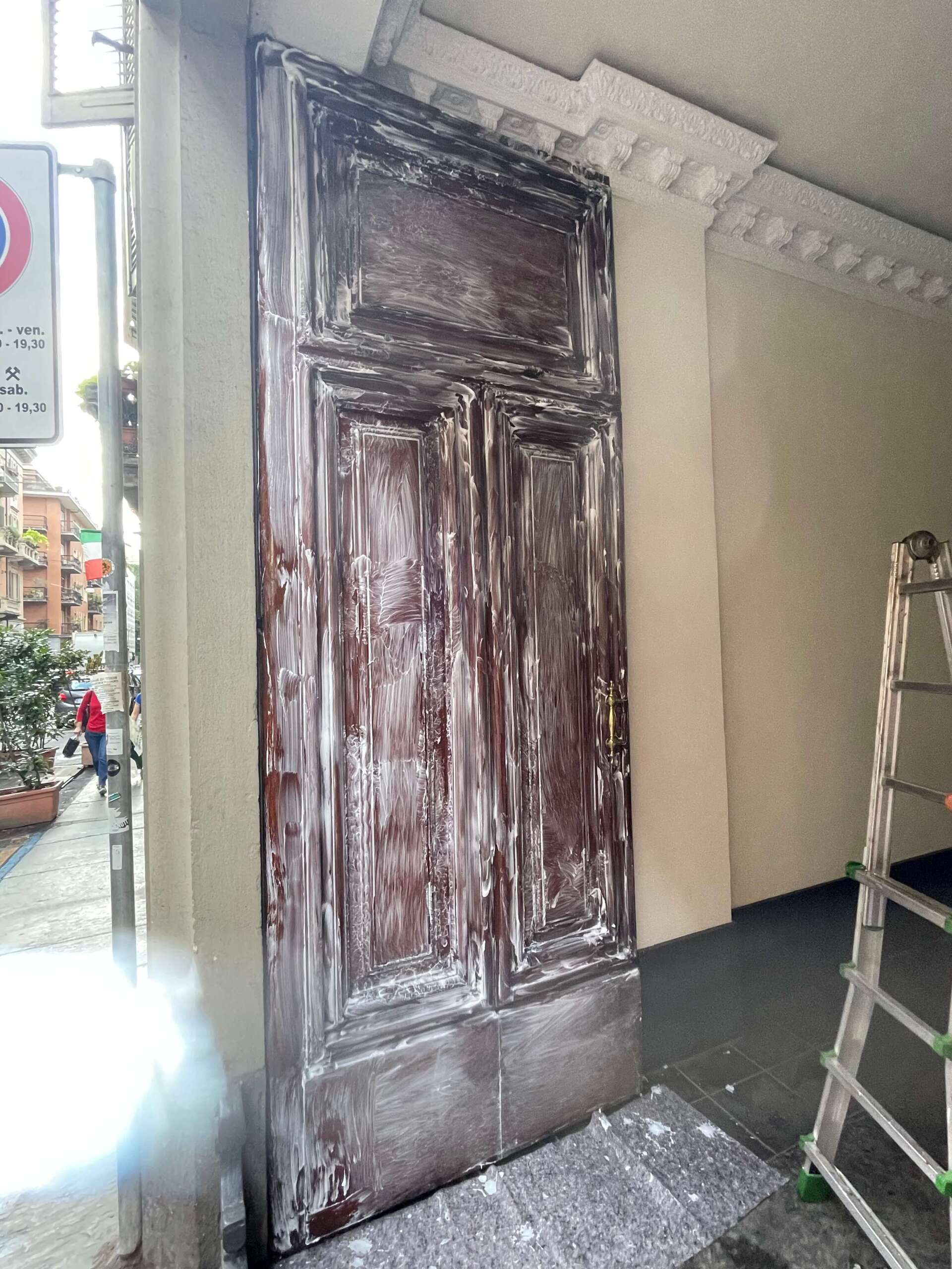 Una porta alta e scura in legno sotto una sporgenza decorativa, parzialmente ricoperta di residui di vernice bianca, con una scala nelle vicinanze.