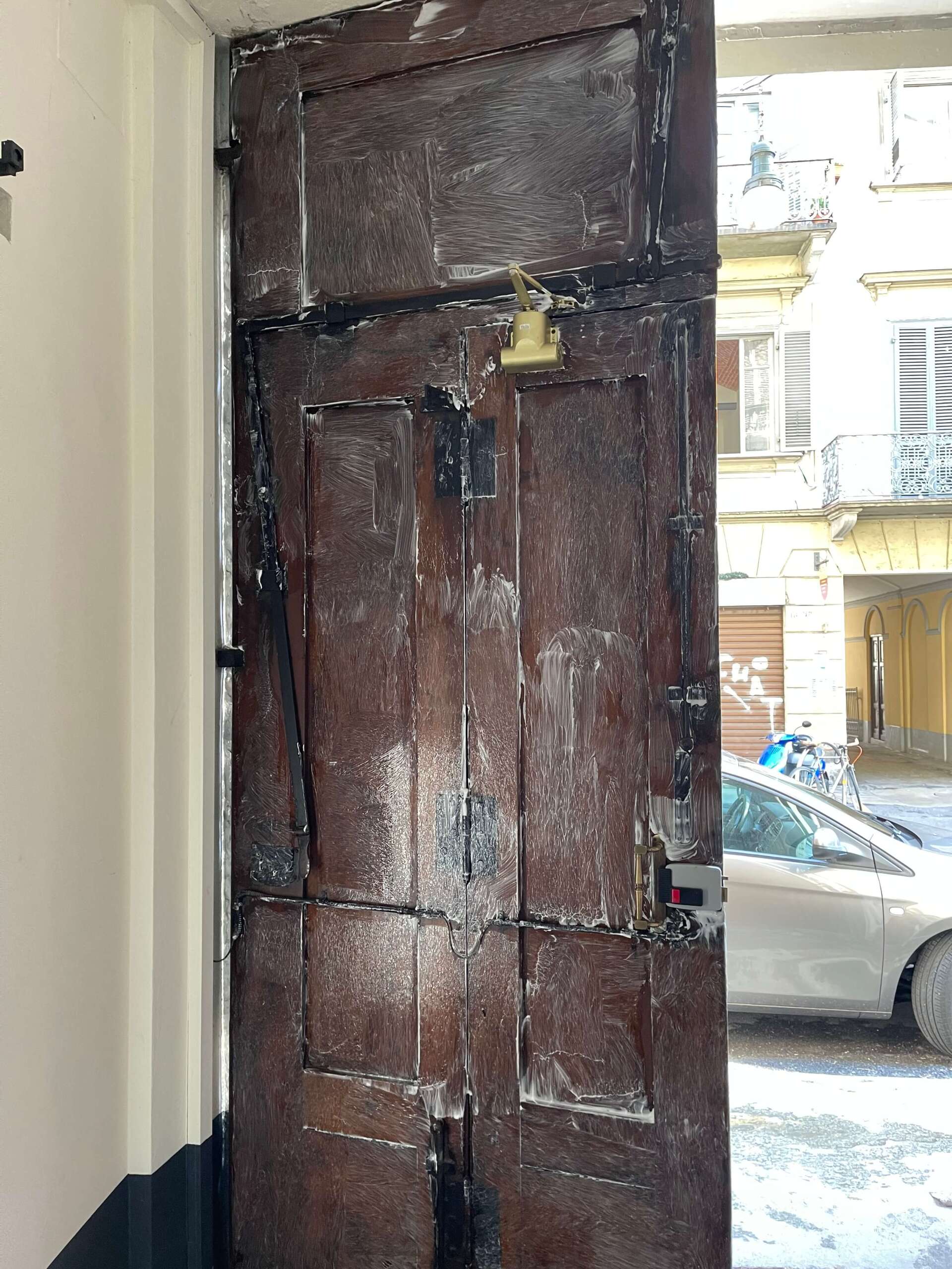 Una porta di legno marrone scuro, usurata dalle intemperie e con la vernice scrostata, vista dall'interno di un portone che guarda verso una strada cittadina.
