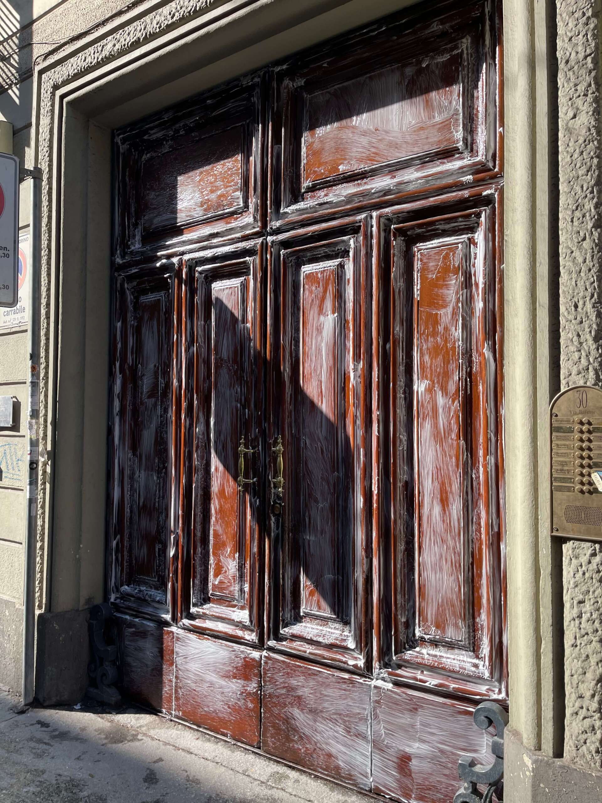 Un paio di alte porte doppie in legno invecchiato con vernice marrone scrostata, inserite in una cornice di pietra con una piccola targa segnaletica.