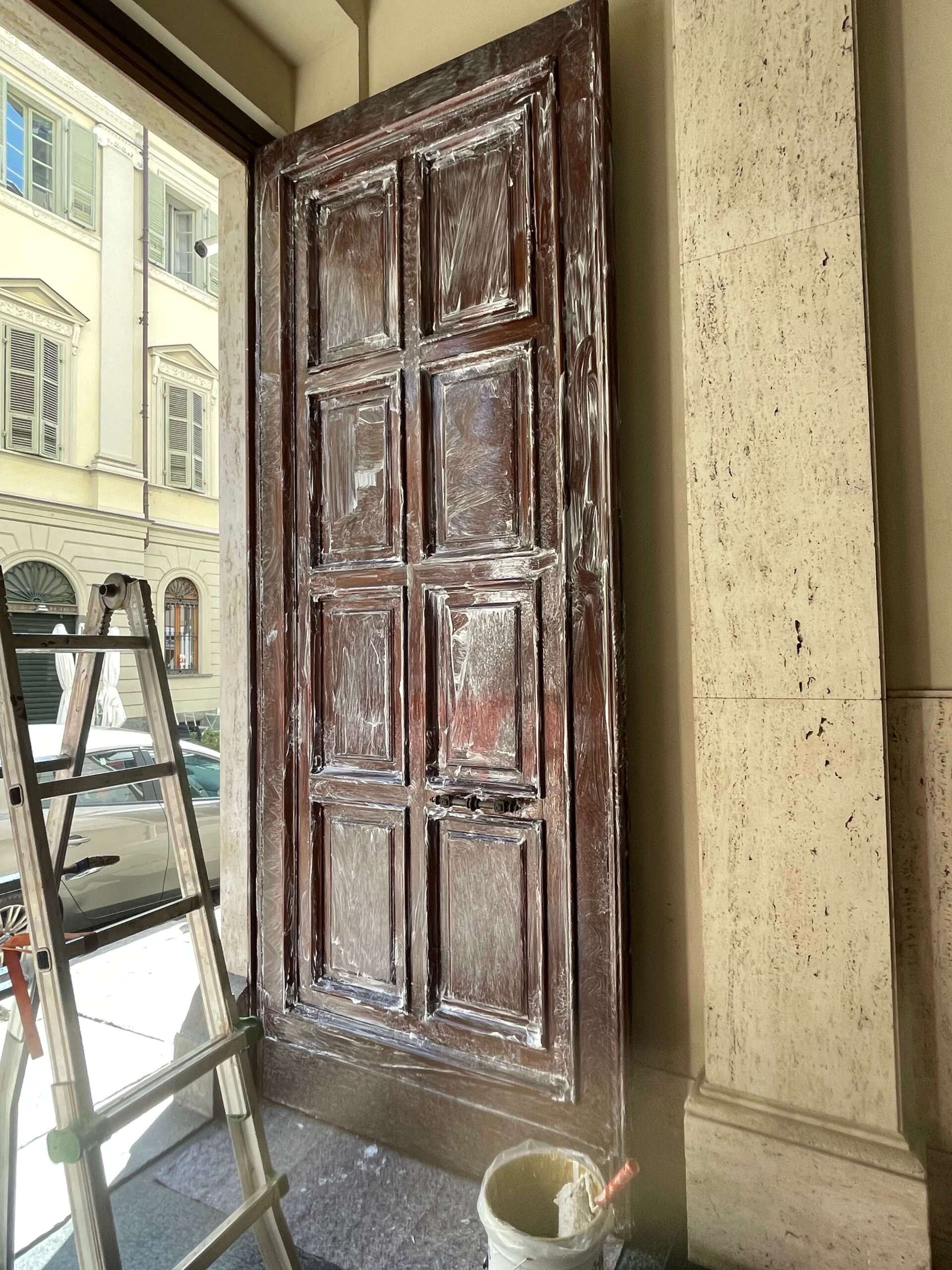 Un'alta porta di legno marrone con pannelli, situata in un vano accanto a una scala e a un secchio di vernice.