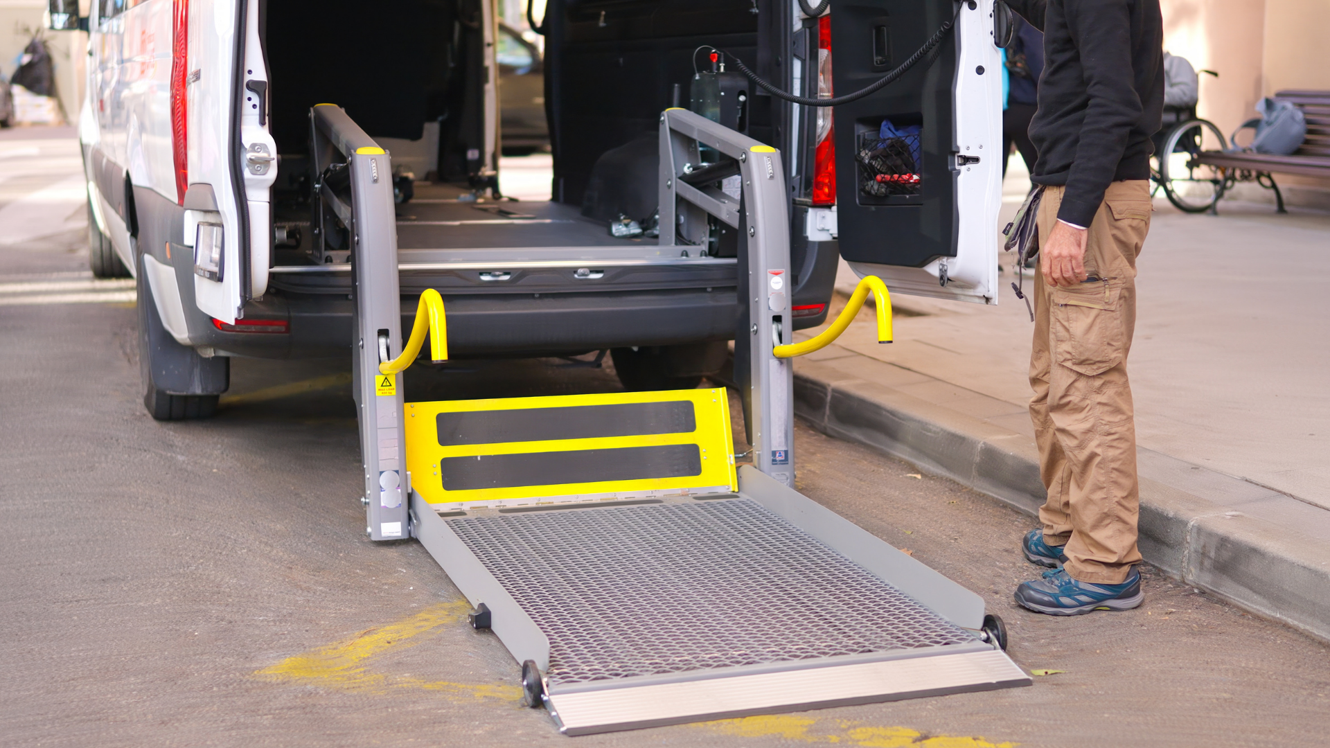A person stands by a van with a deployed hydraulic wheelchair lift extended to ground level on a paved surface.
