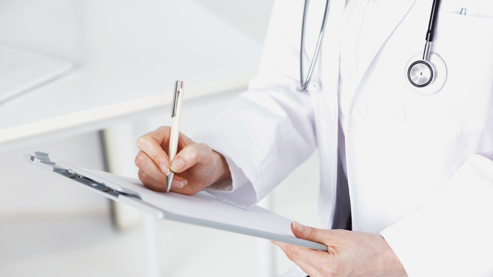 A medical professional in a white coat with a stethoscope around their neck writing on a clipboard.
