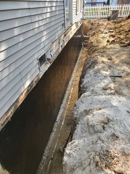 Home foundation trench beside gray siding, exposing basement wall and dirt excavation path