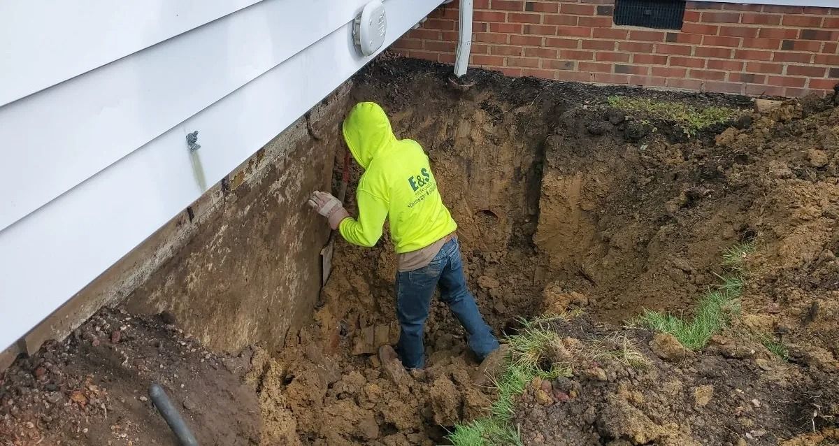 Worker in neon green jacket digging soil beside a house foundation in a muddy yard