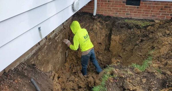 Worker in neon green jacket digging soil beside a house foundation in a muddy yard