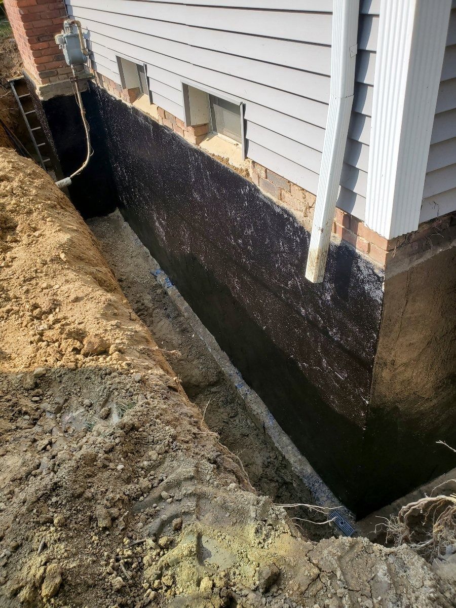Exterior house foundation with black waterproofing and a dug trench beside the siding