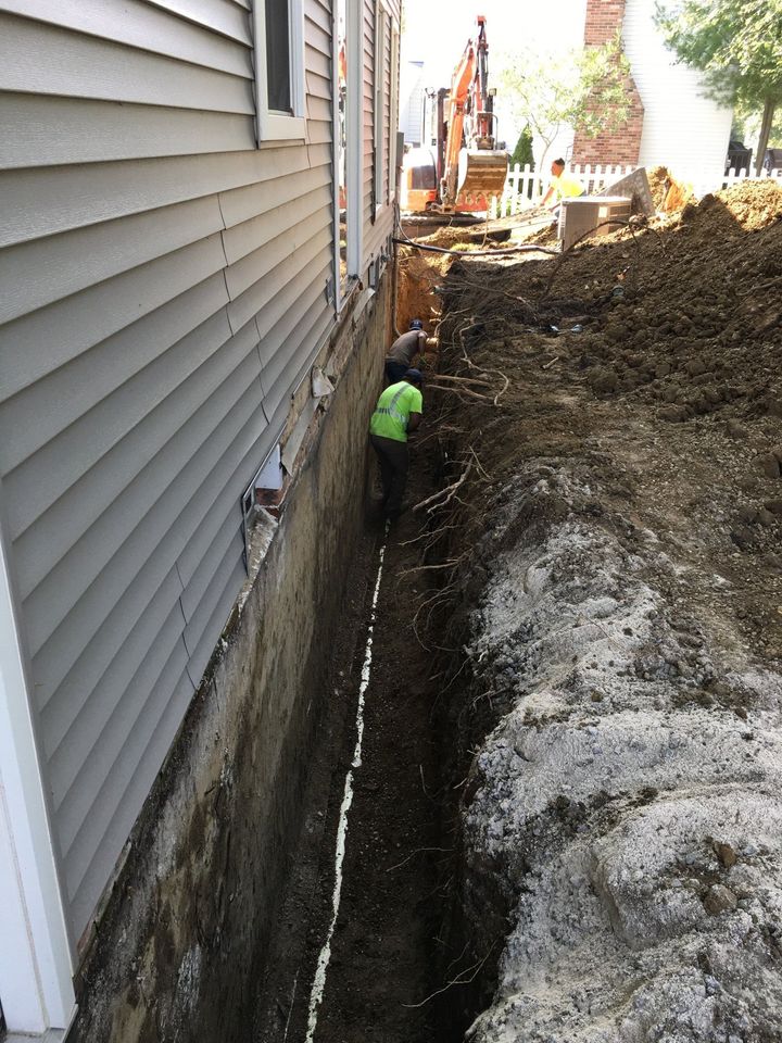 Narrow trench beside house siding with a worker in a neon vest digging near an excavator.
