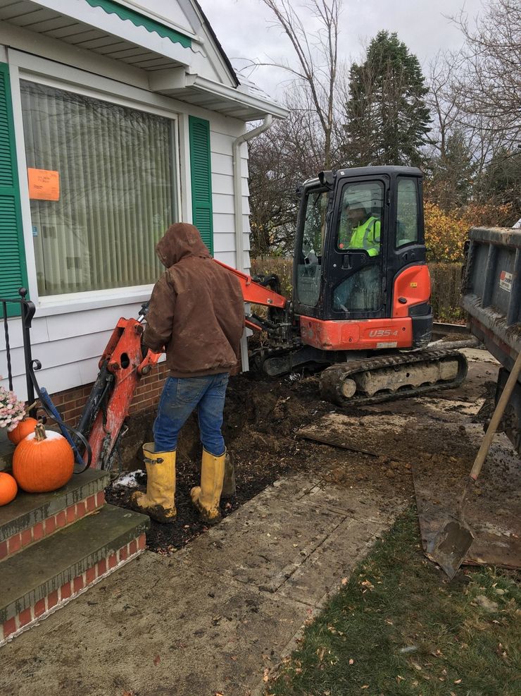 Person using a mini excavator beside a house, with pumpkins on the porch and a muddy yard