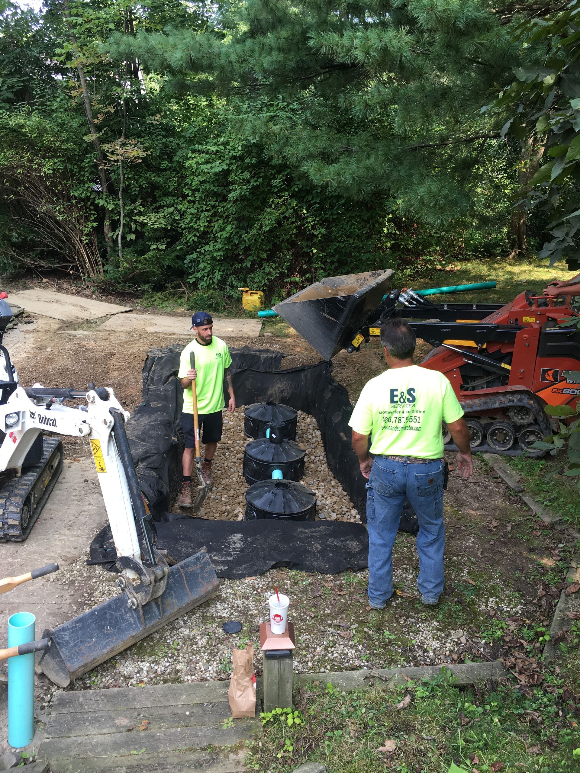 Workers inspecting a large collapsed hole in a wooded area beside heavy equipment and a red truck.