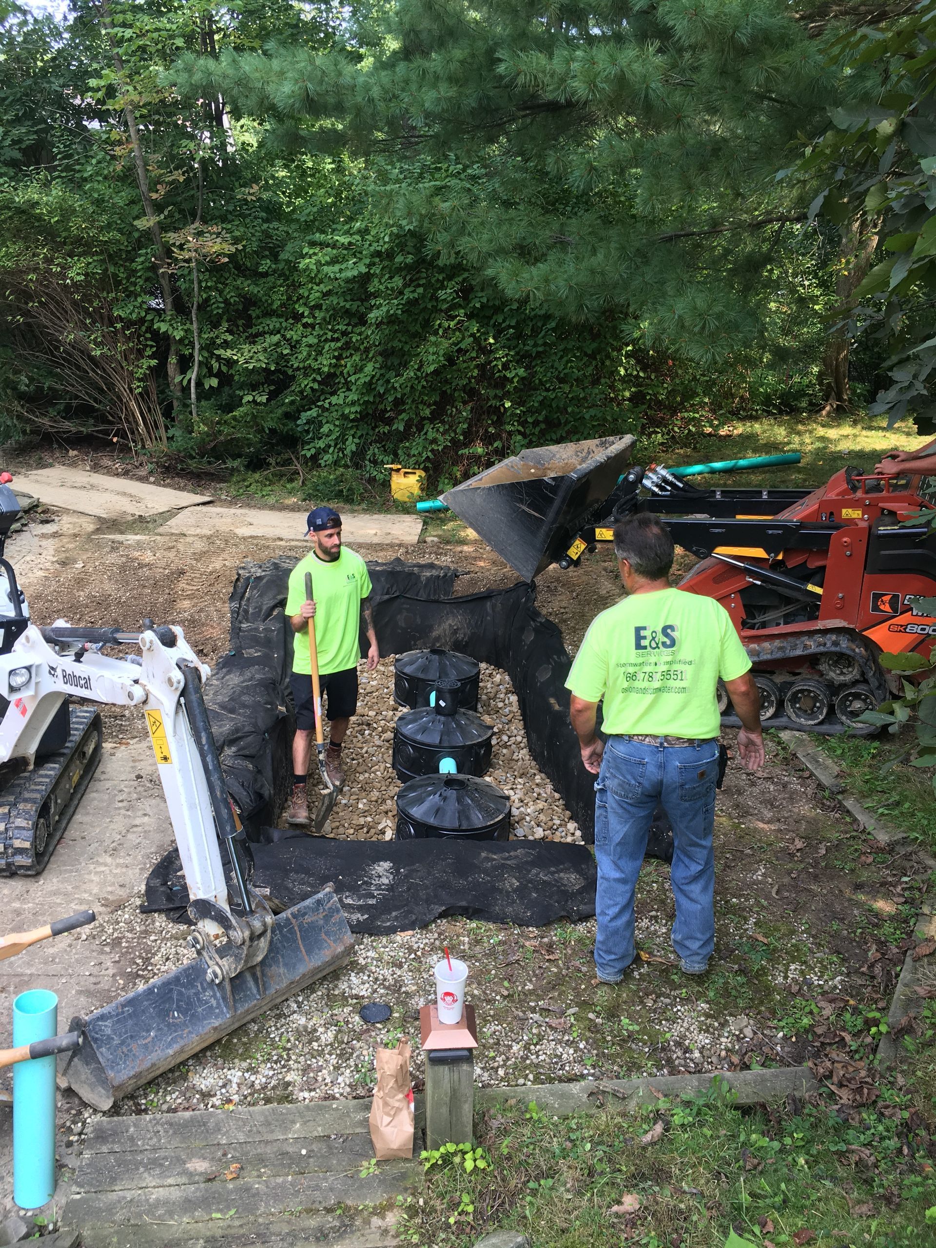 Workers inspect a mud-covered black car at a wooded accident scene with heavy machinery nearby.