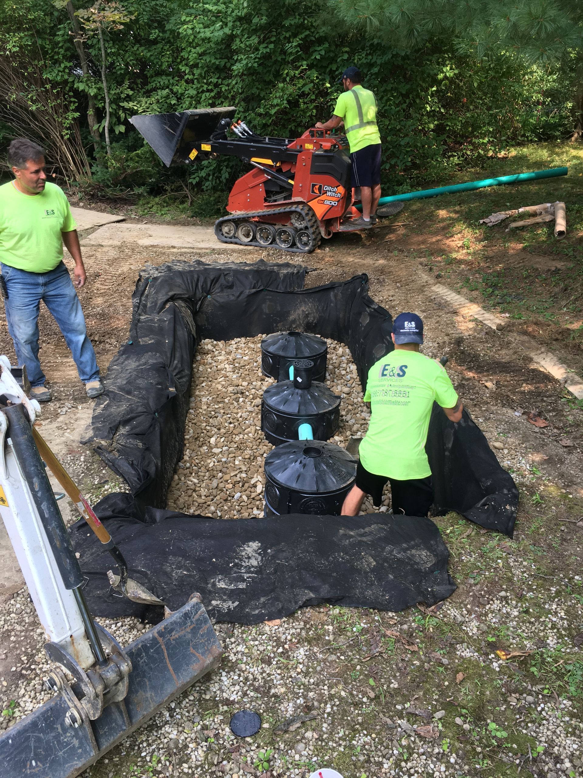 Workers install a black-lined trench with drainage pipes beside a wooded path