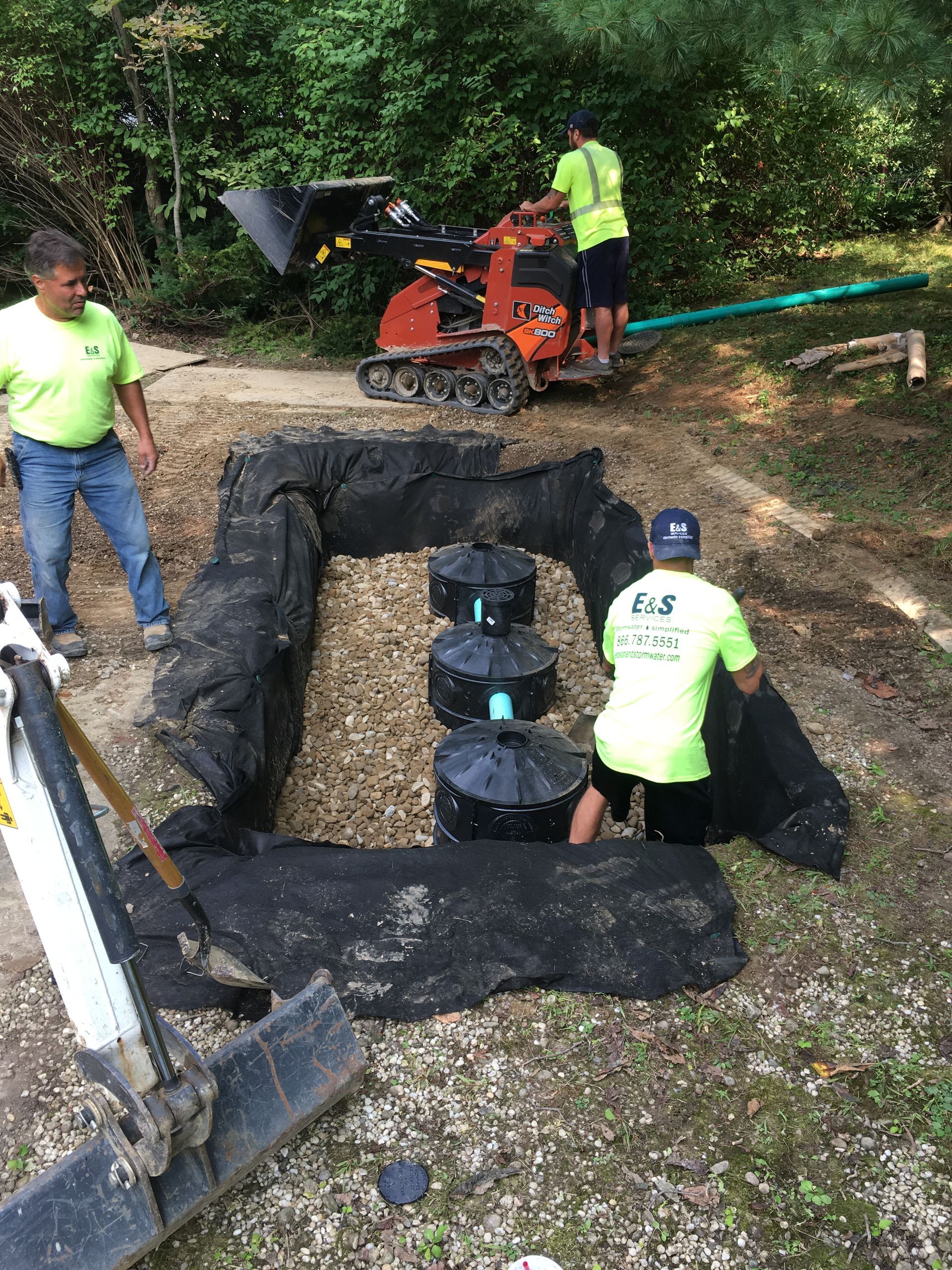 Workers installing black landscape fabric around a small outdoor path with a compact machine nearby.