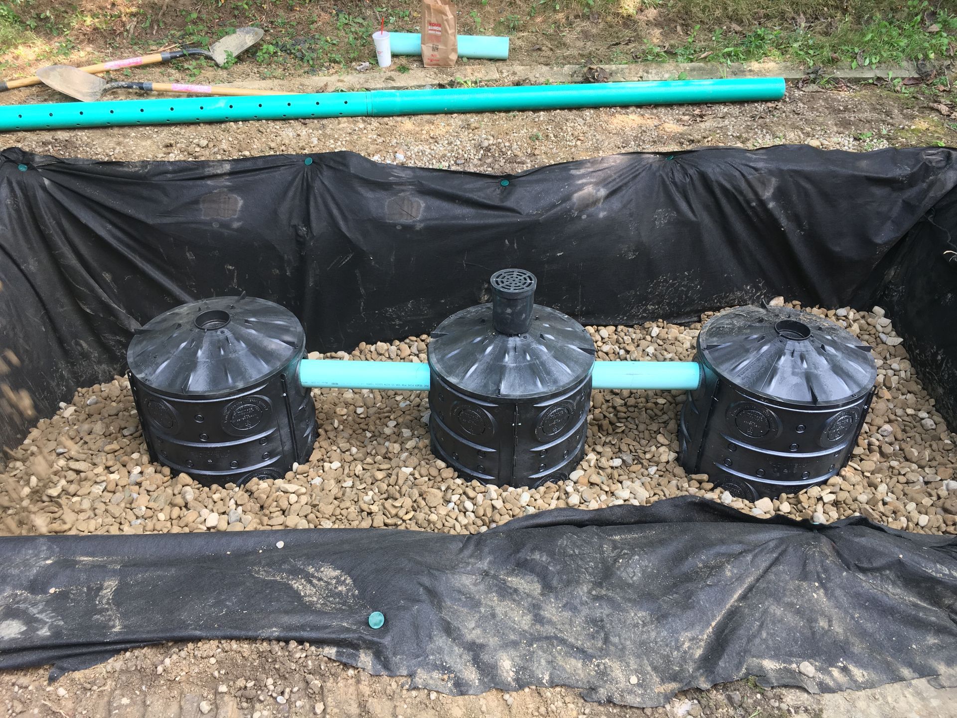 Three black buried barrels linked by blue pipes in a lined trench with gravel around them.