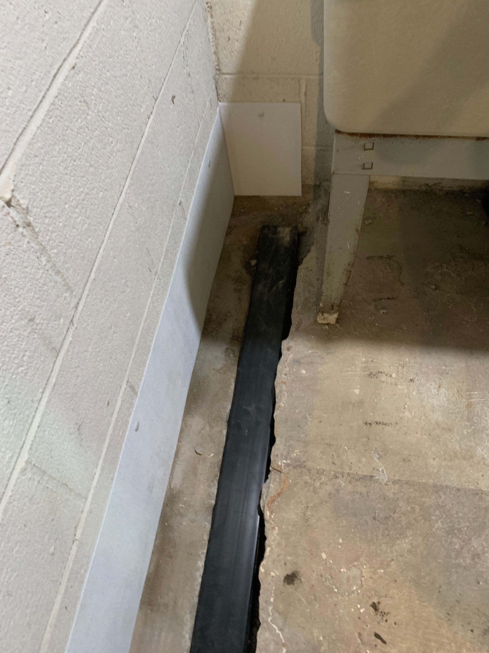 Narrow floor gap along a cinder block wall, filled with black sealant in a concrete room