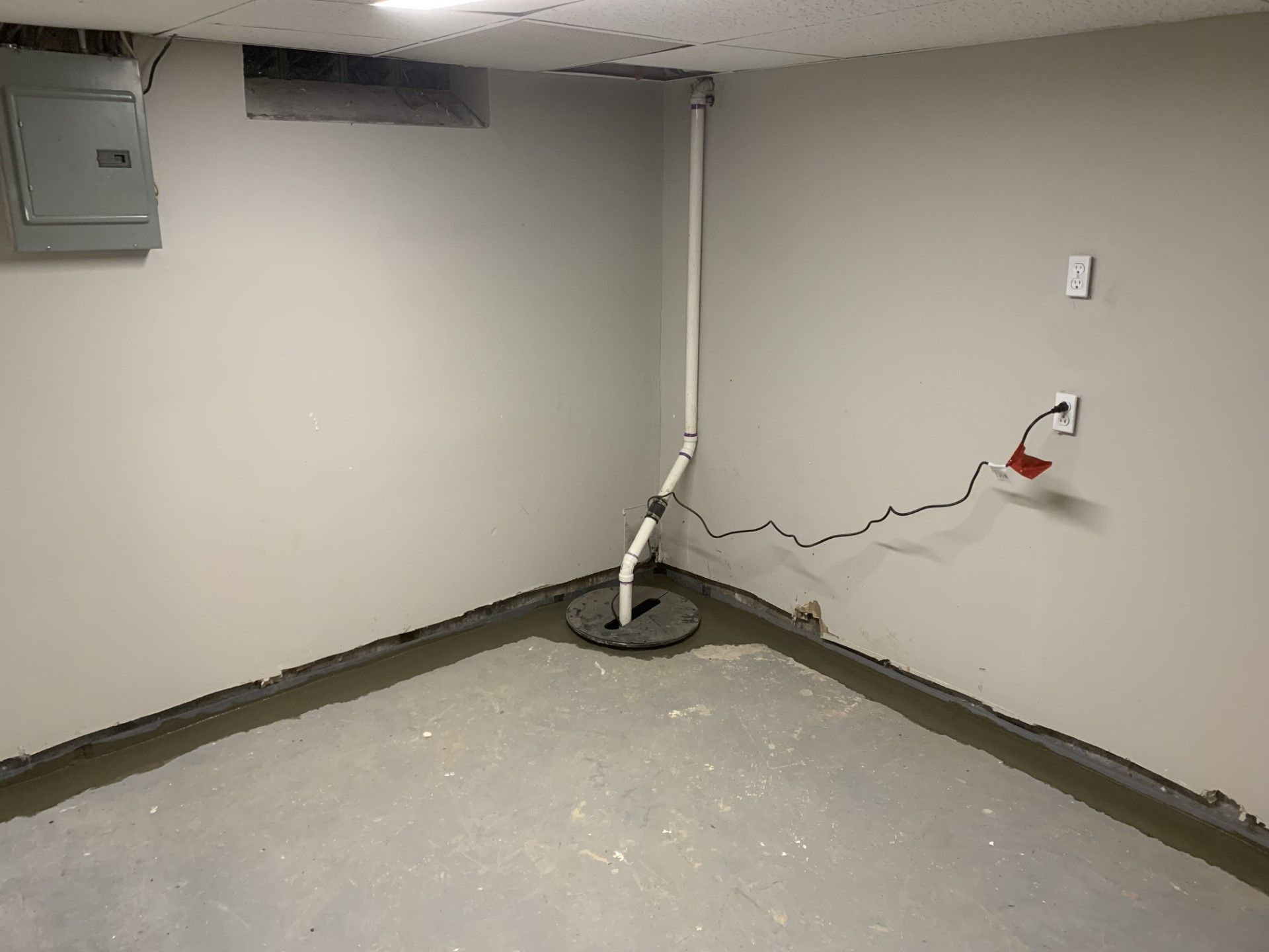 Empty utility room with gray walls, exposed piping, electrical box, and a damaged wall outlet with loose wires