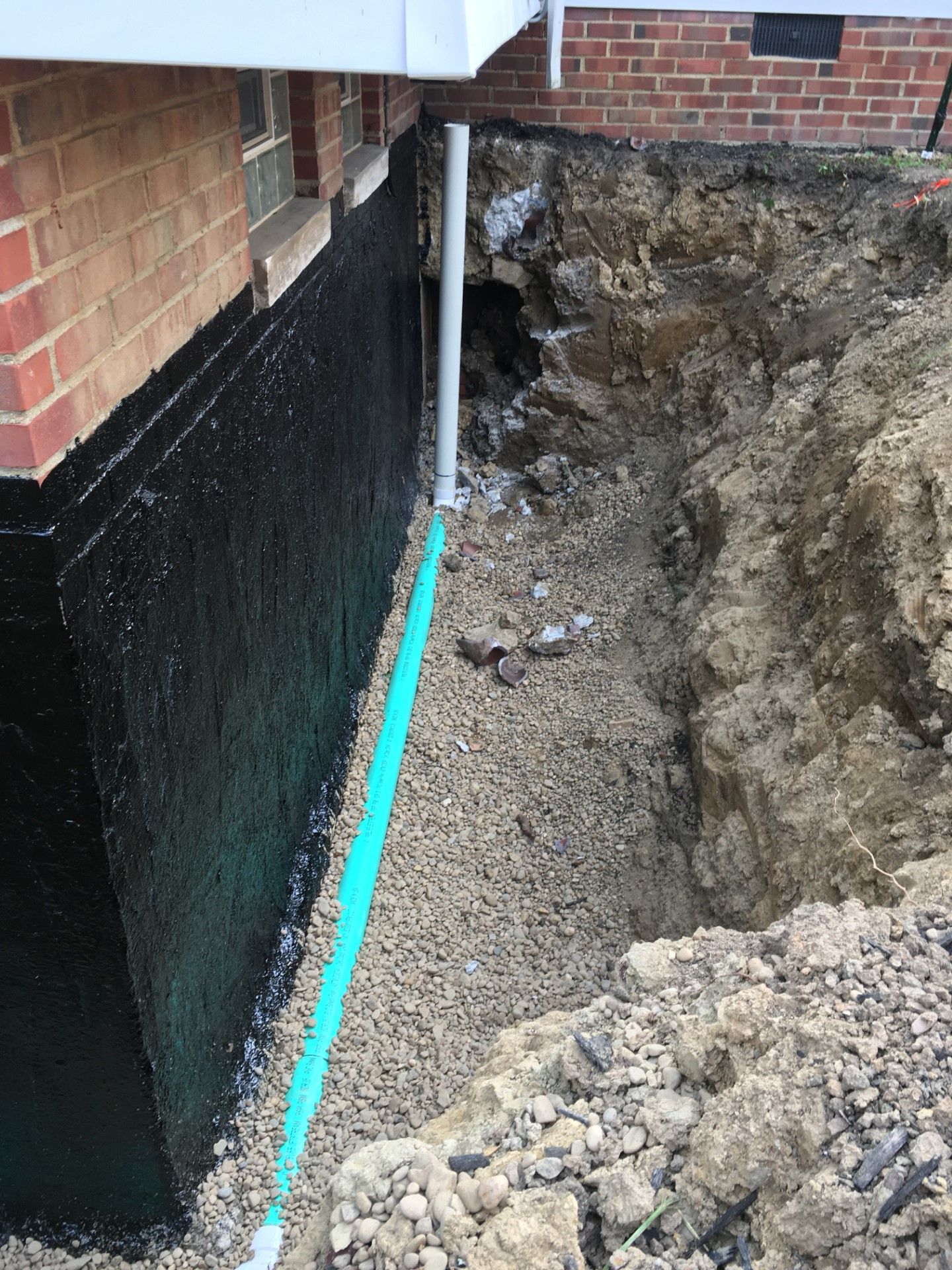 Basement foundation excavation beside brick house with black waterproofing and gravel trench