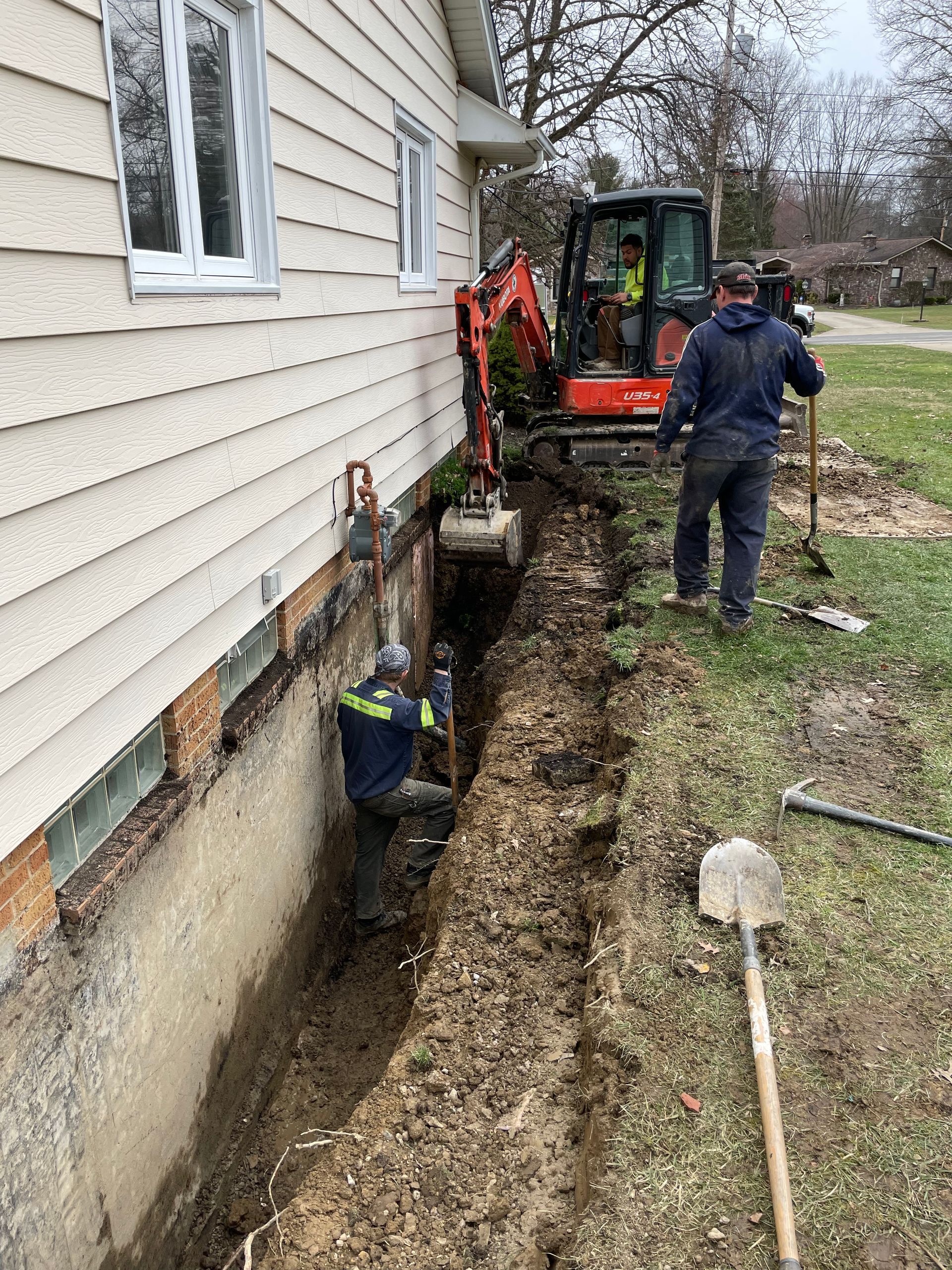 Backhoe digging a trench beside a house while a worker stands nearby.