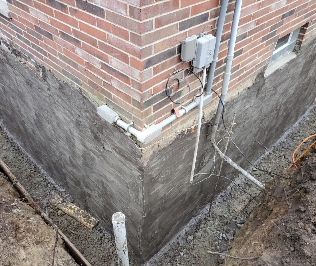 Foundation excavation around a brick house corner with gray waterproofed walls and exposed utility lines