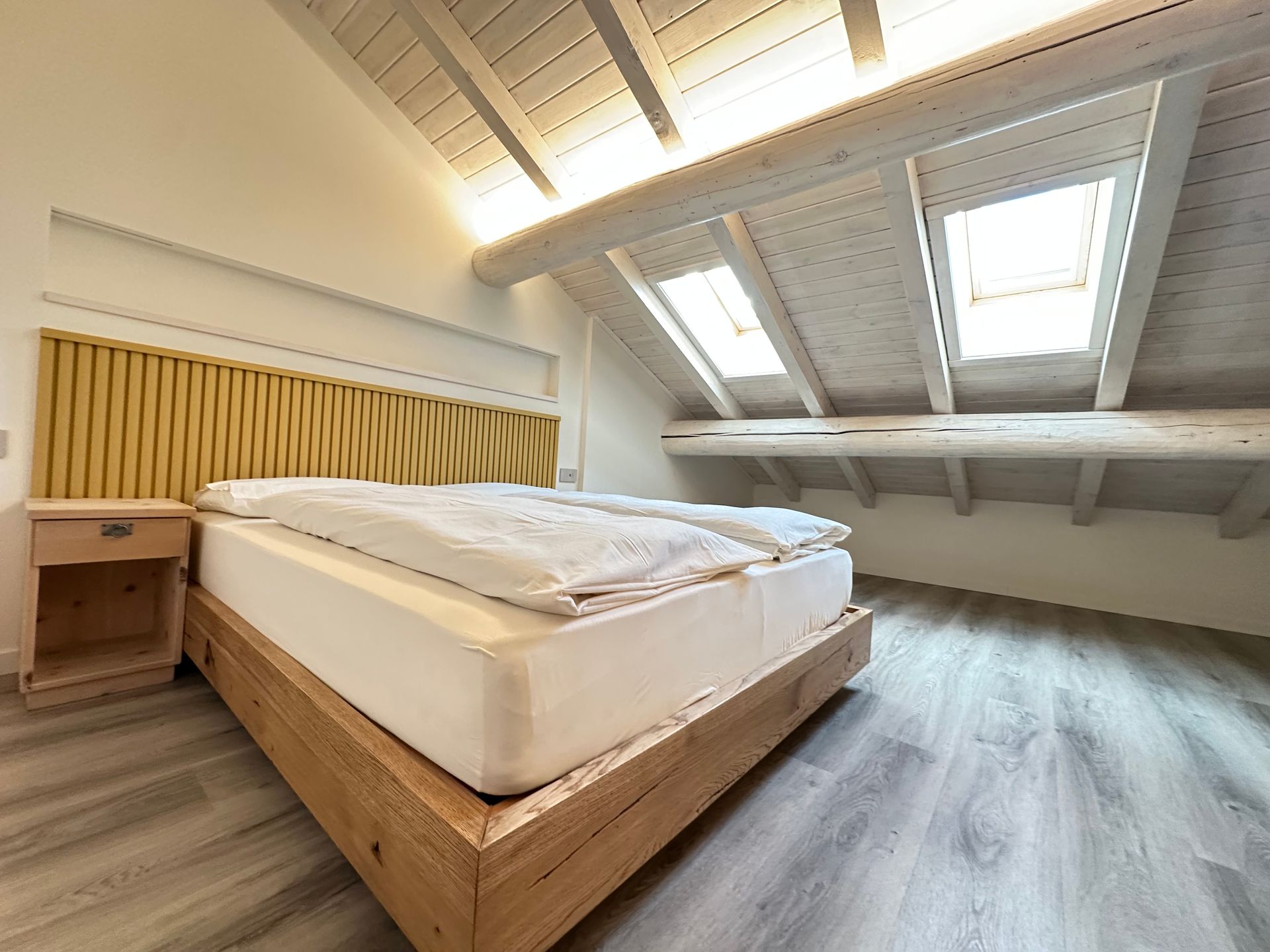 Camera da letto con letto, comodino e lucernari. Legno chiaro, pareti bianche e testiera gialla.