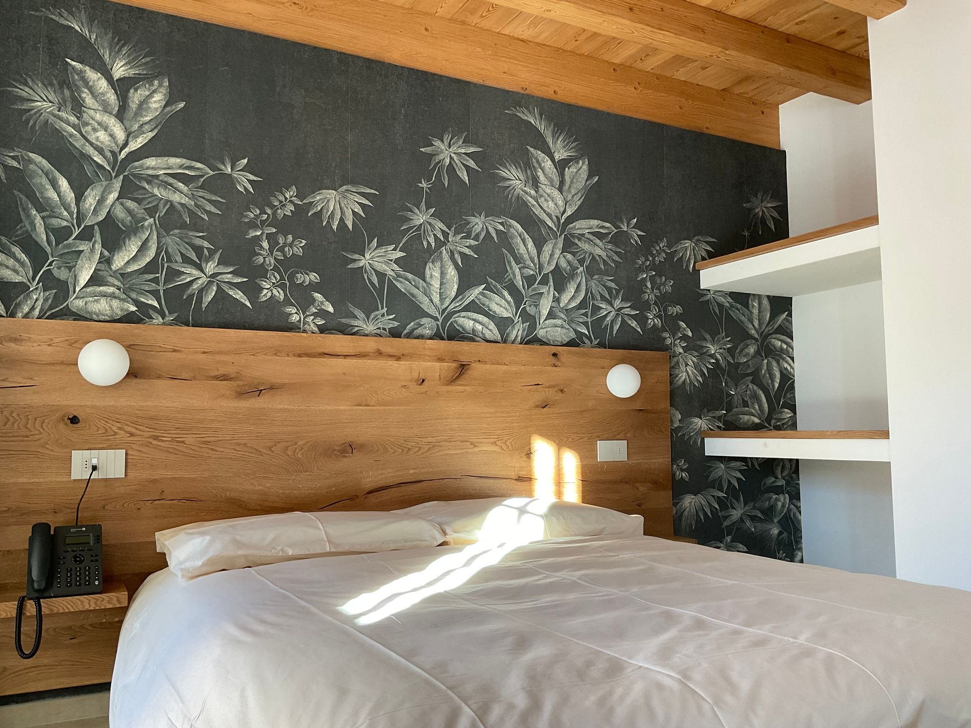 Camera da letto con ampia testiera in legno, biancheria da letto bianca, carta da parati floreale e scaffali incorporati.