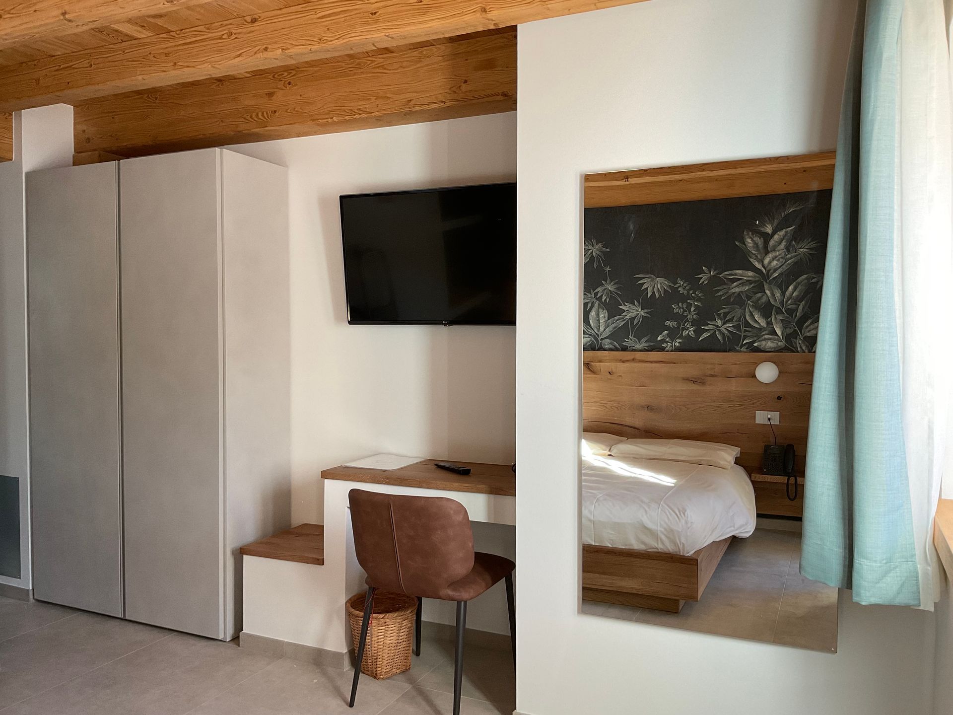 Camera da letto con letto, scrivania, armadio e specchio. Pareti bianche, soffitto in legno e una tenda azzurra.
