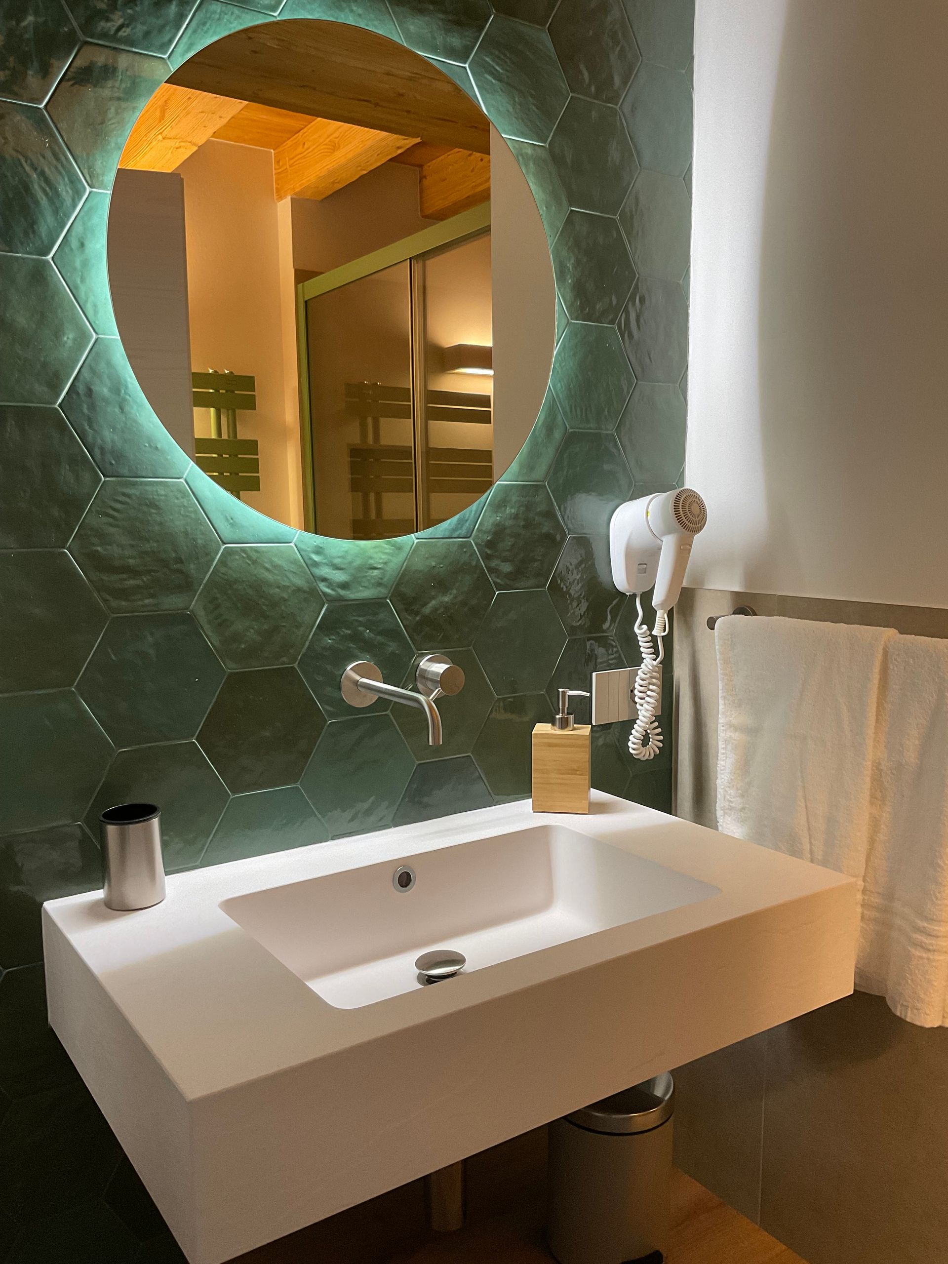 Bagno con lavandino bianco, specchio rotondo illuminato, parete con piastrelle esagonali verdi.