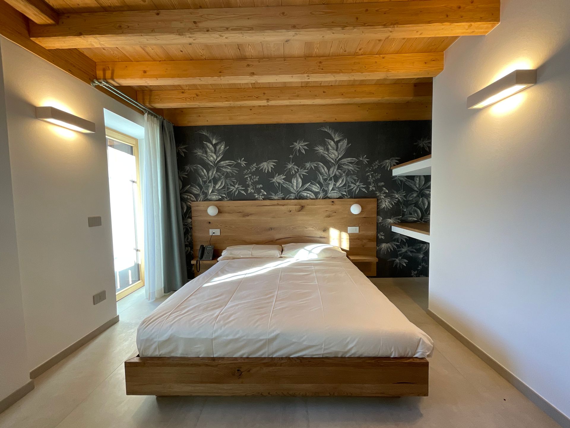 Camera da letto con letto in legno, testiera e travi a vista; carta da parati floreale scura e due applique.