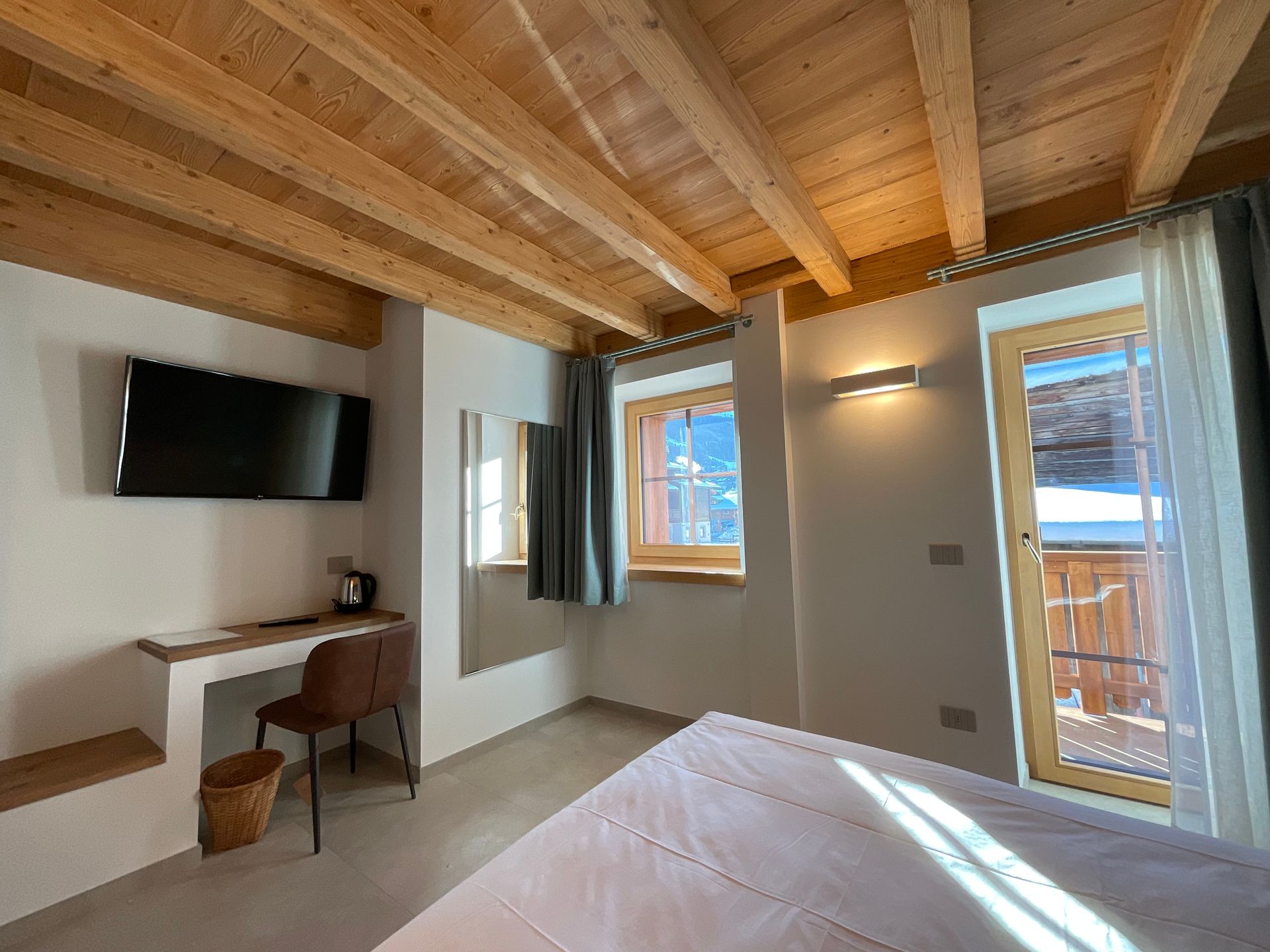 Camera da letto con soffitto in legno, TV, scrivania e finestre che si affacciano su un balcone innevato.