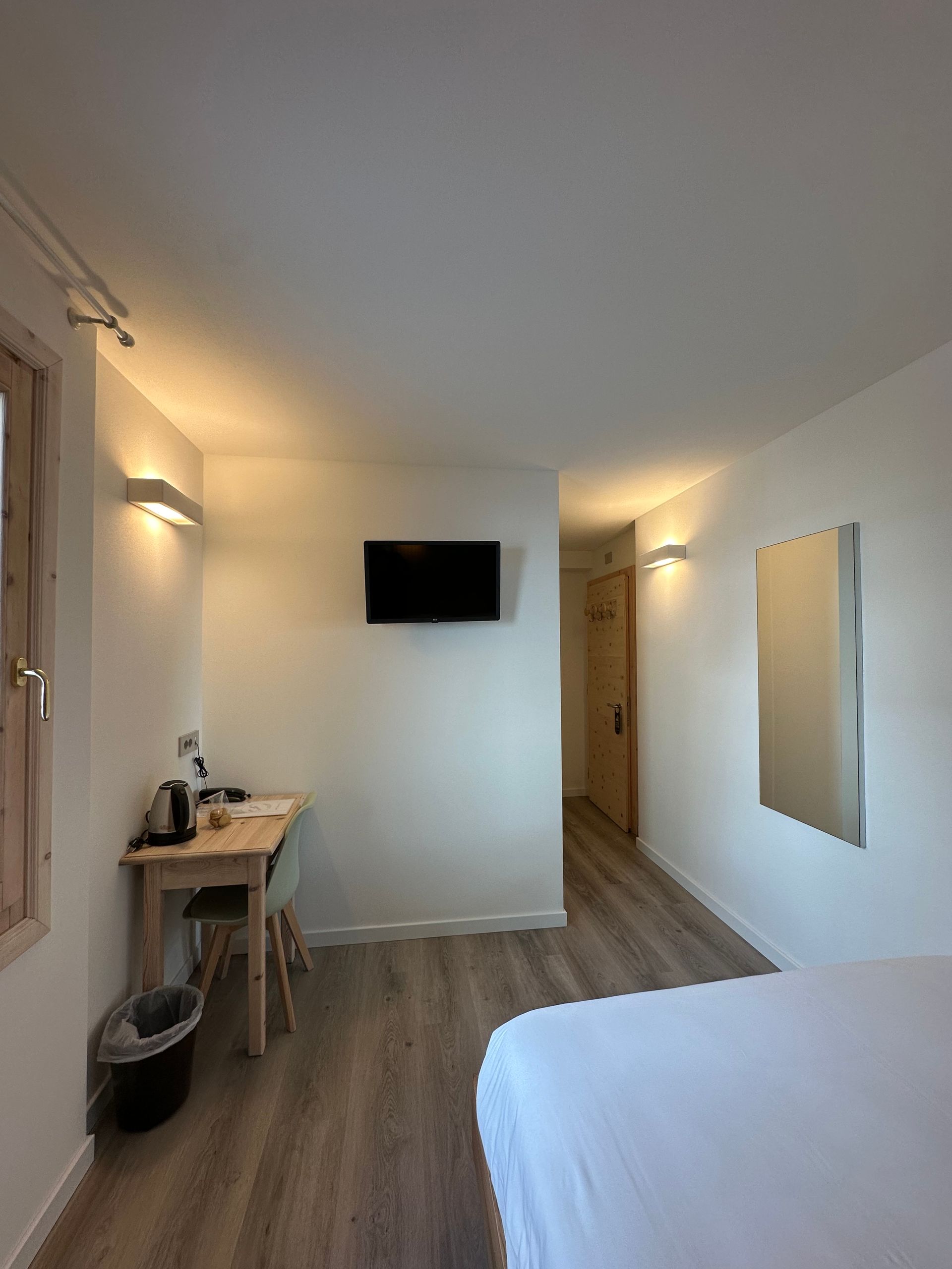 Camera d'albergo con letto, scrivania, TV, specchio e corridoio. Pareti bianche, pavimento in legno.