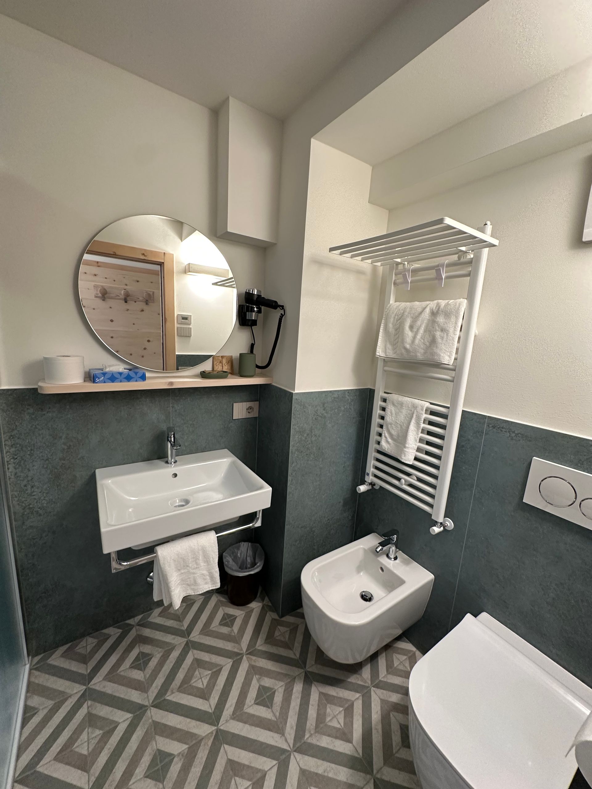 Piccolo bagno con lavandino, WC e bidet bianchi. Pareti piastrellate color verde acqua. Specchio rotondo e portasciugamani.