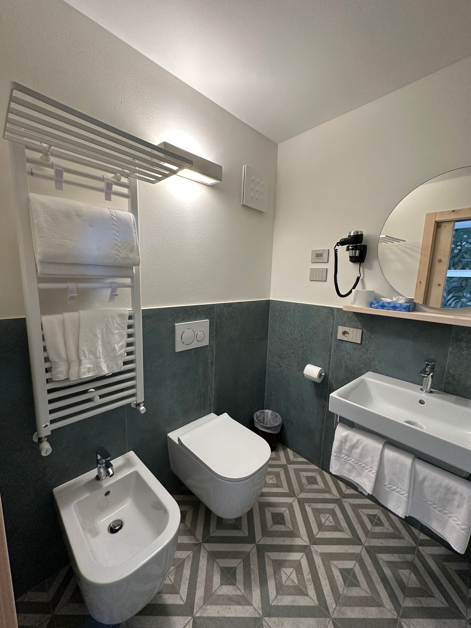 Bagno con WC, bidet, lavandino e scaldasalviette. Colori grigio e bianco, pavimento a motivi.
