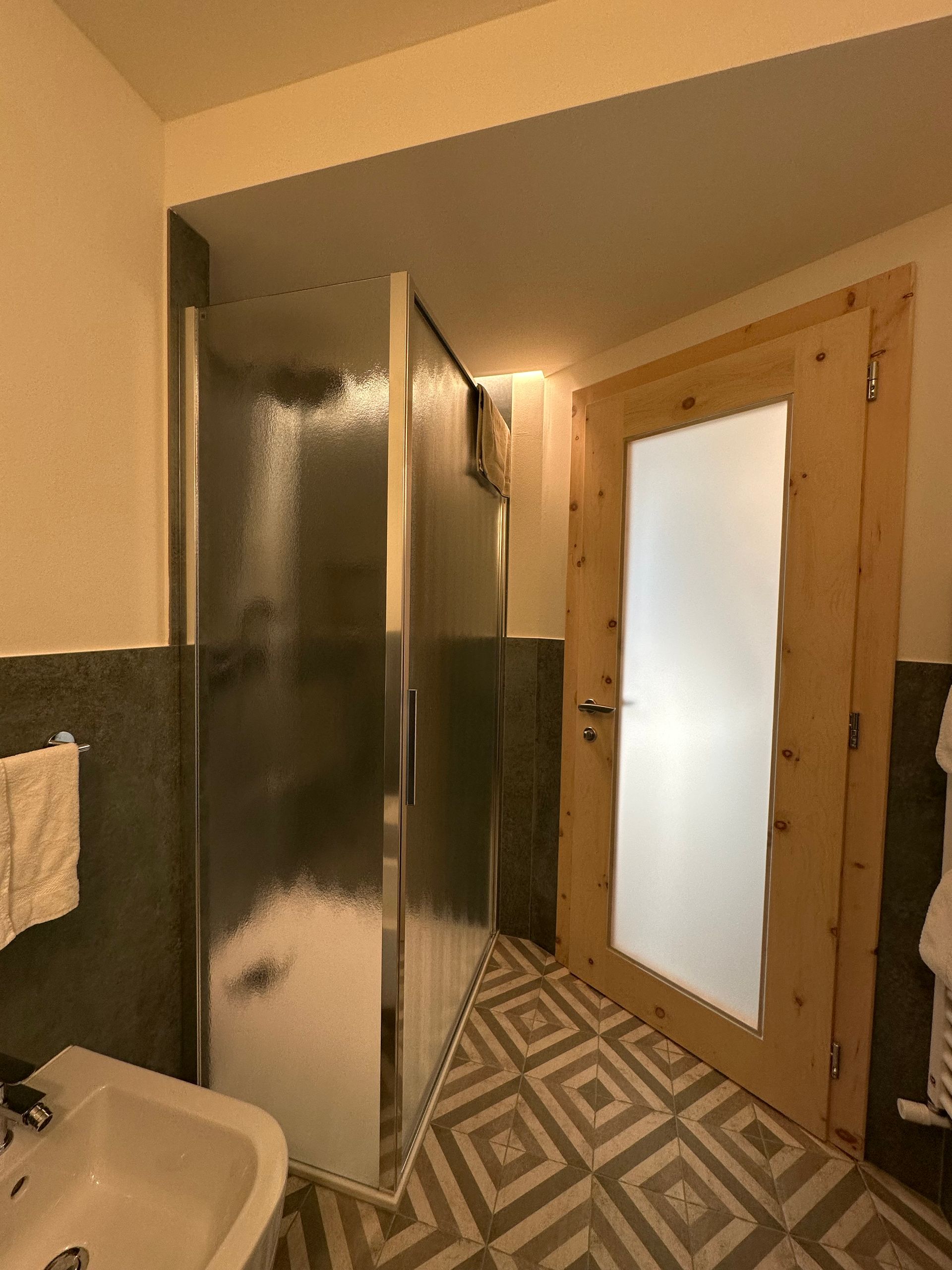 Bagno con box doccia e porta in legno con vetro smerigliato. Pavimento piastrellato e pareti con dettagli verde scuro.