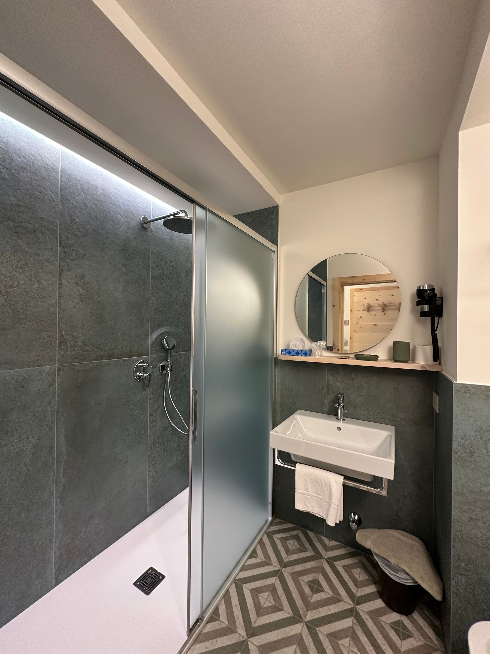 Bagno con doccia piastrellata grigio scuro, porta in vetro smerigliato, lavandino bianco, specchio rotondo e pavimento decorato.