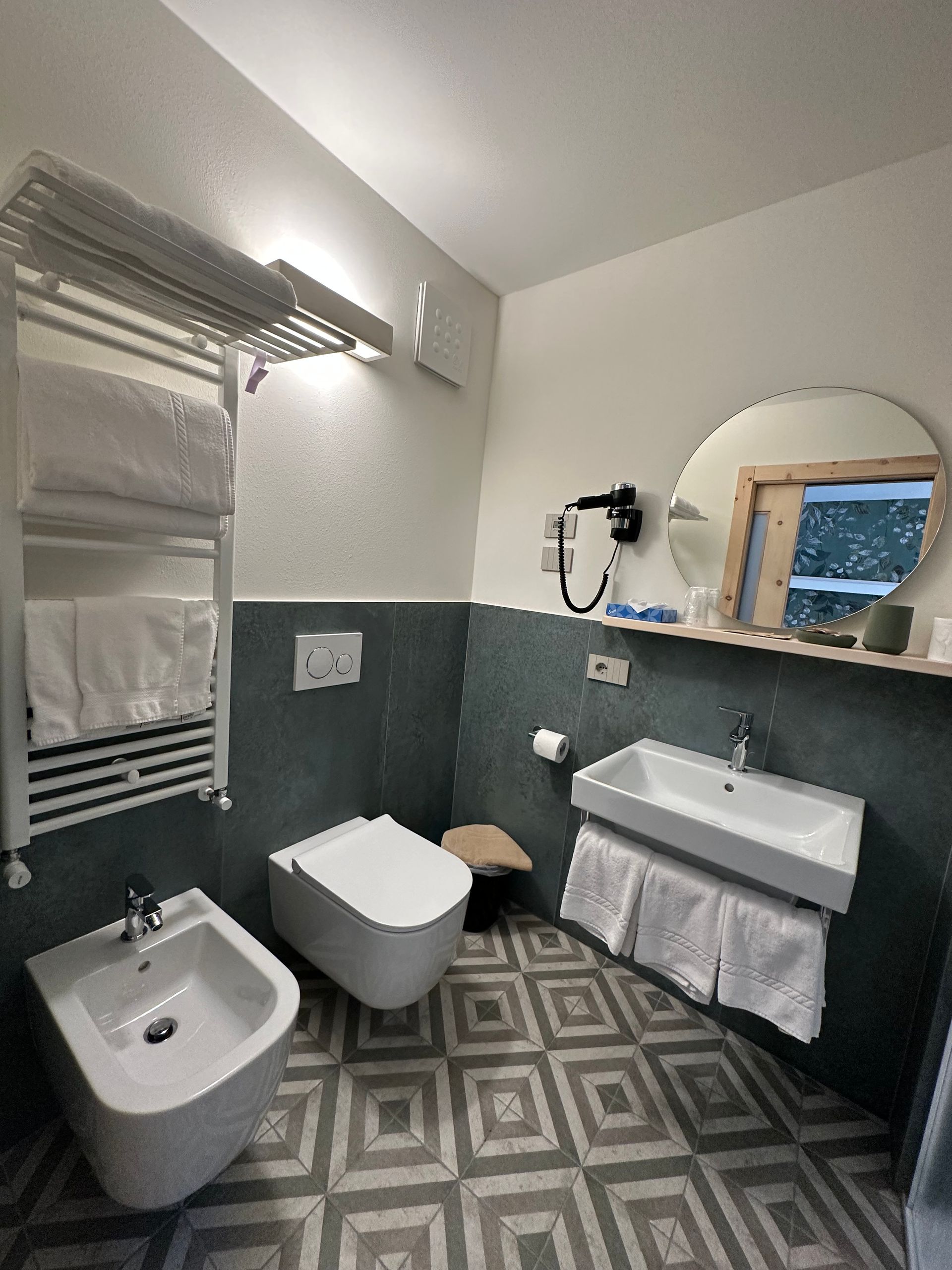 Bagno con WC, bidet, lavandino, specchio e portasciugamani. Presenta pavimenti in piastrelle grigie e bianche e pareti con dettagli blu-verdi.