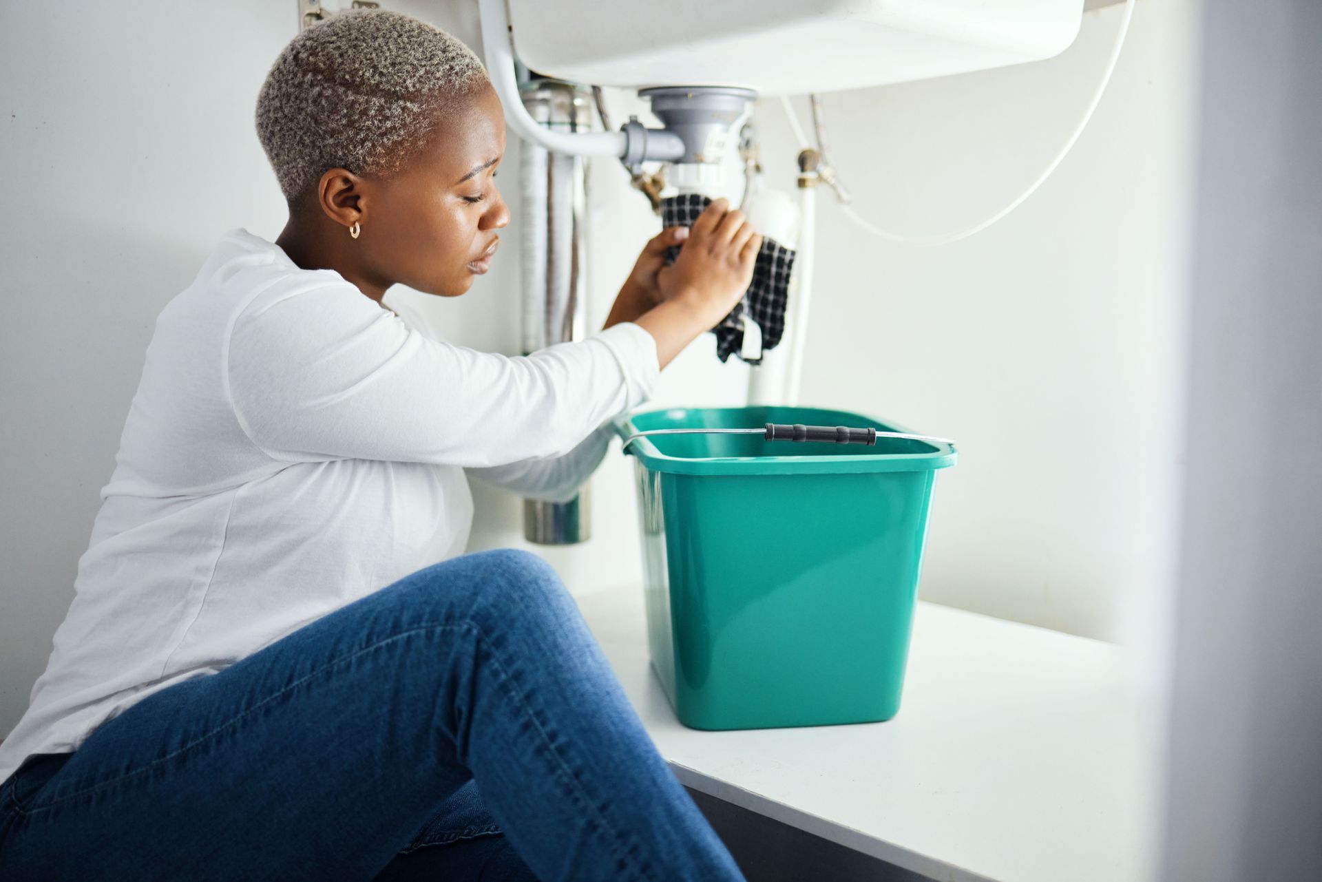 A worried woman is with a cloth and a bucket, stopping a burst pipe, while waiting for assistance.