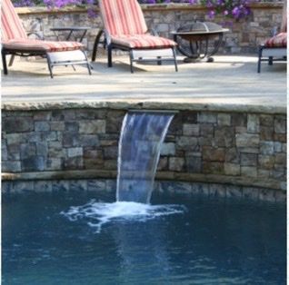 Water cascades from a stone wall into a pool, with lounge chairs and fire pit visible.