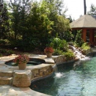 Pool area with stone waterfall, hot tub, and gazebo surrounded by greenery.