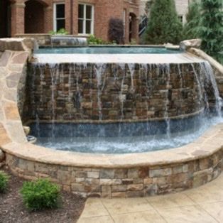 Water cascading over a stone wall into a curved pool, set in front of a brick building.