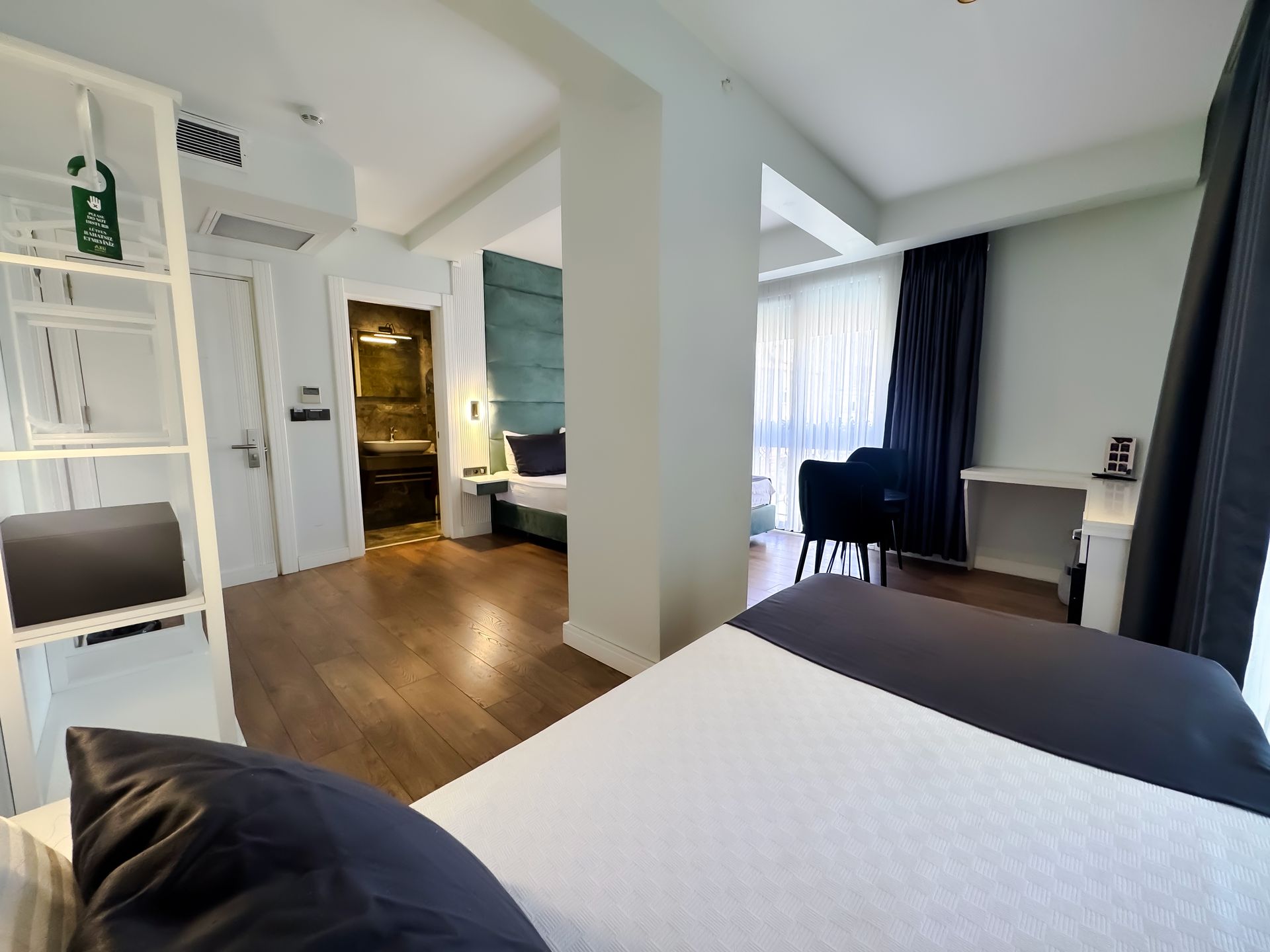 Modern hotel room with bed, desk, dark wood floor, and large window with sheer curtains