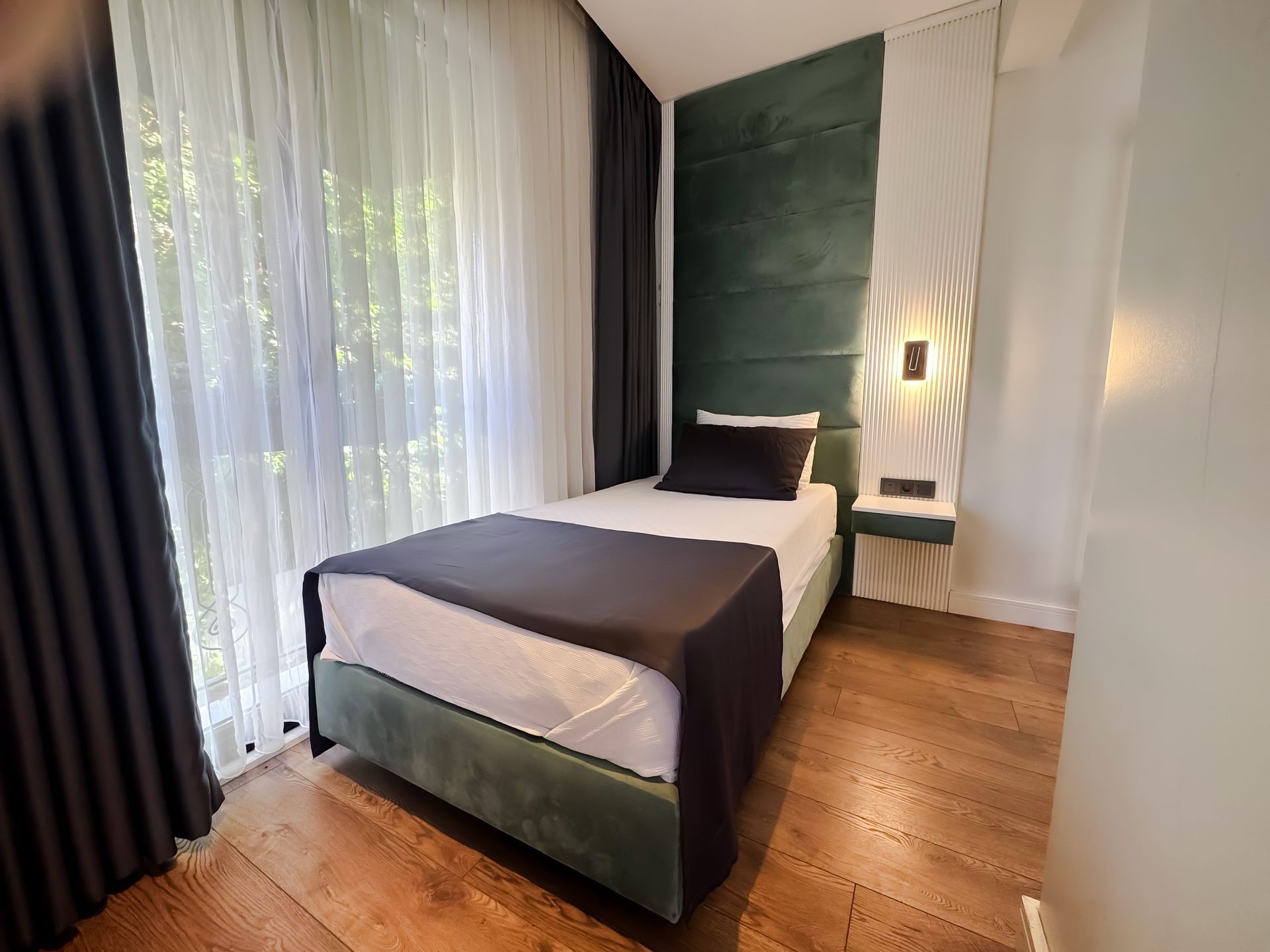 Small bedroom with single bed, dark curtains, wooden floor, and green accent wall with bedside light.
