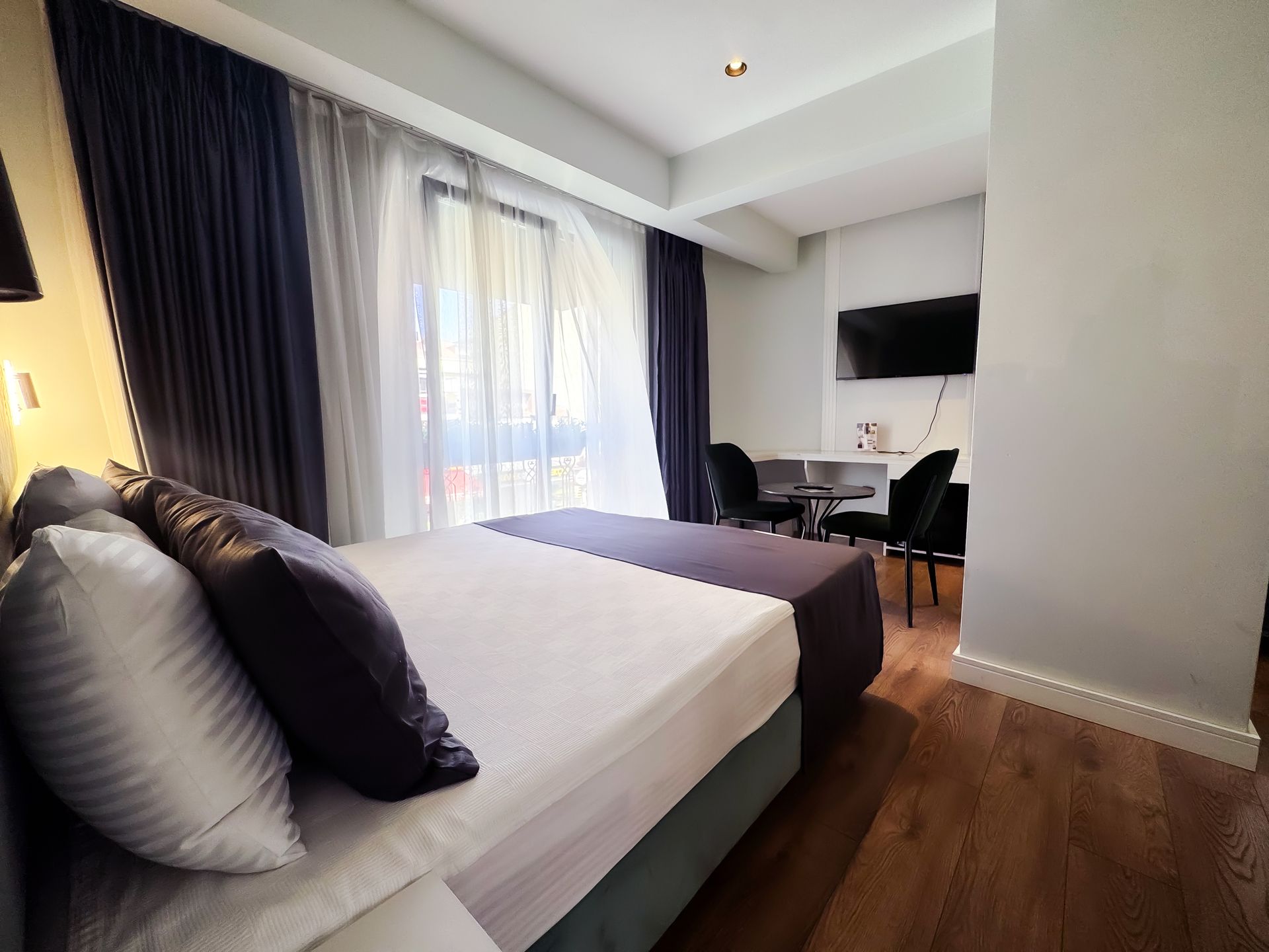 Modern hotel room with bed, dark curtains, desk, and wall-mounted TV near a window