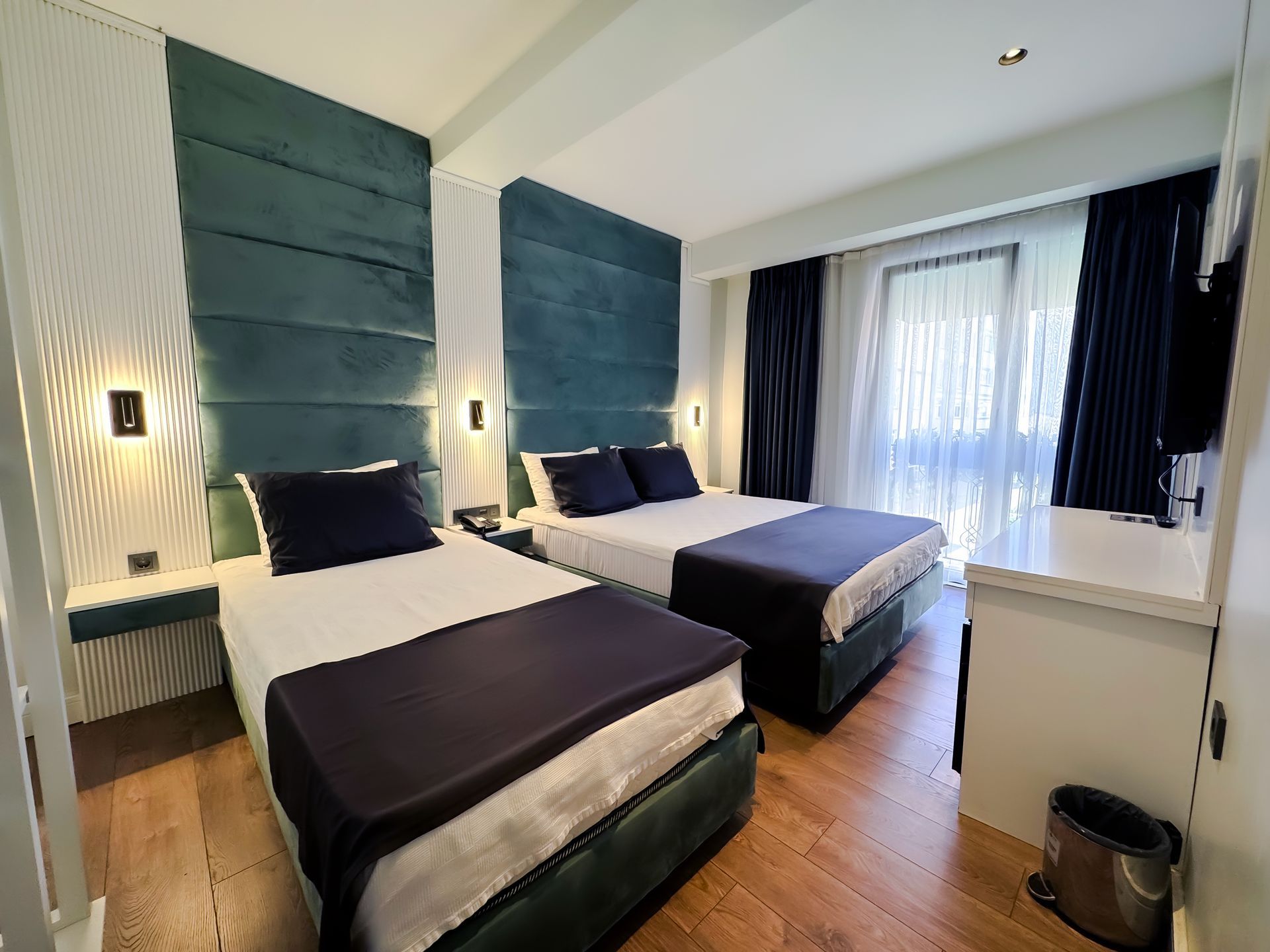 Modern hotel room with two twin beds, blue headboards, and a balcony door with sheer curtains