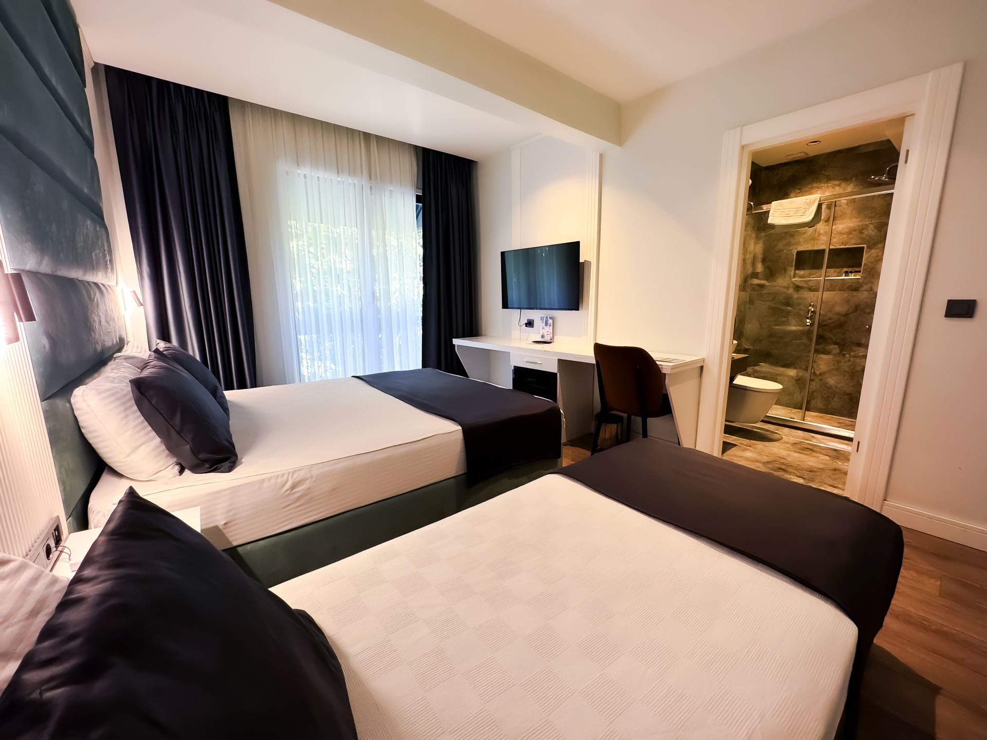 Modern hotel room with two beds, white linens, dark accents, TV, desk, and bathroom door opening on the right