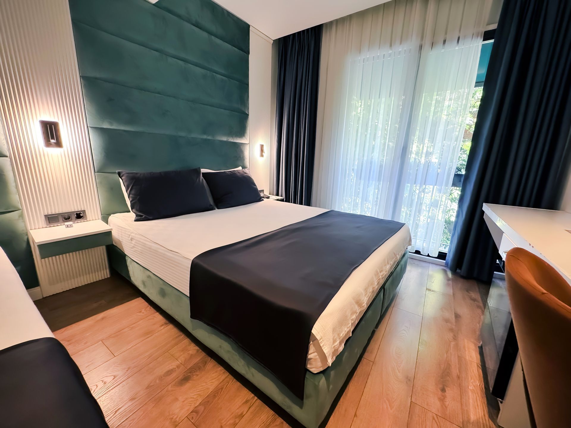 Modern bedroom with teal upholstered bed, dark bedding, wood floor, and large sheer-curtained windows