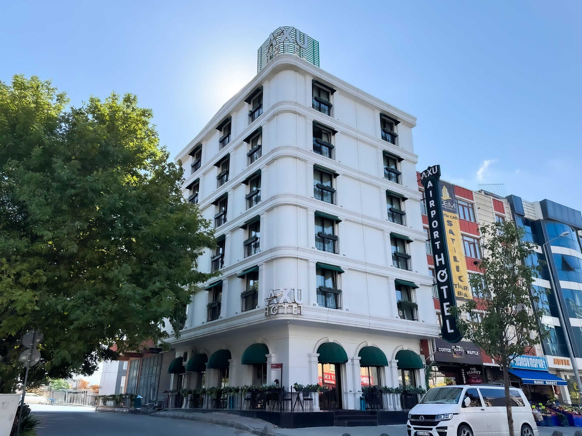 White corner hotel building with arched windows beside a tree-lined street and colorful apartment blocks