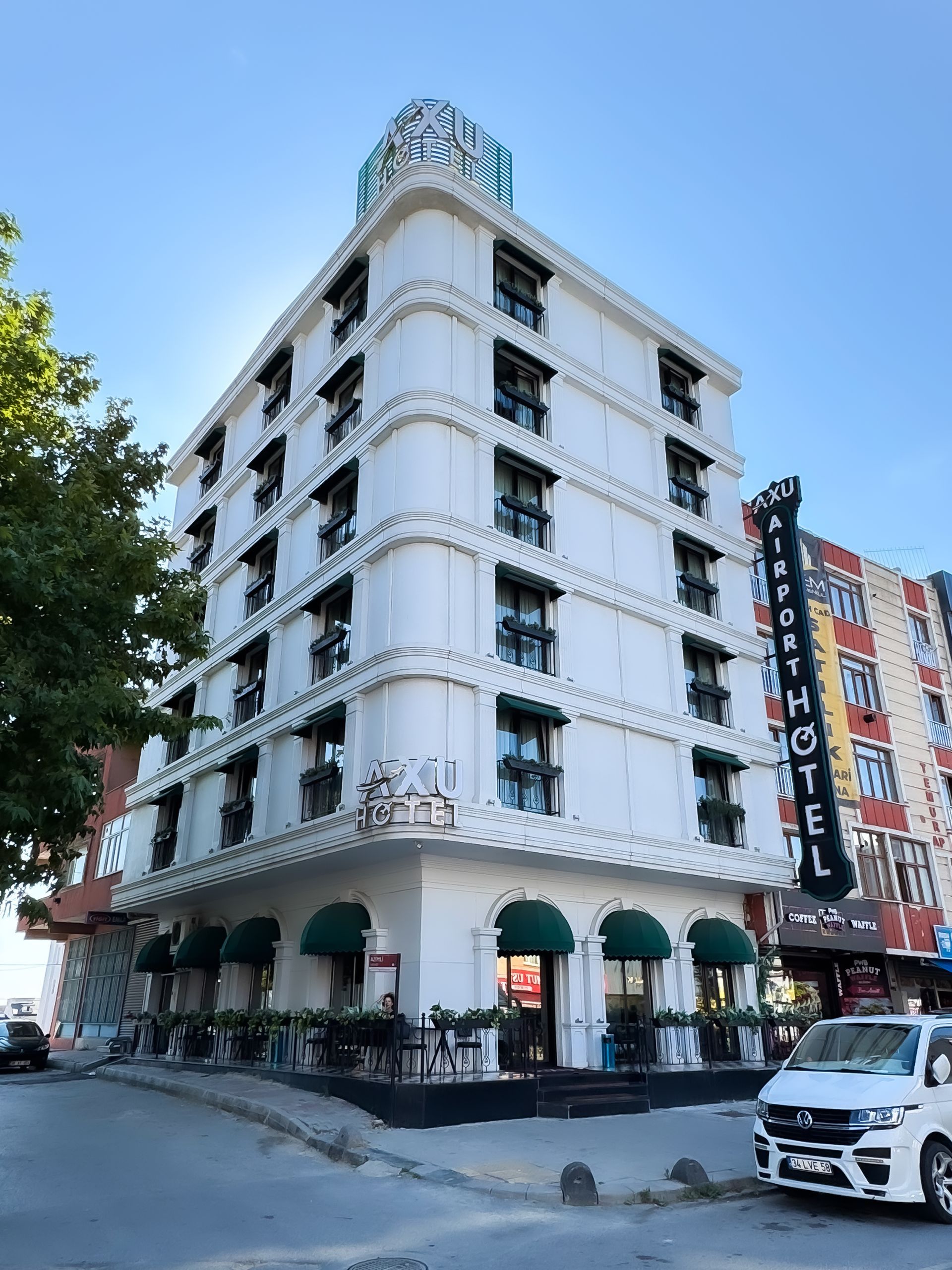White corner hotel building with arched ground floor, balconies, and a vertical neon sign on a city street.