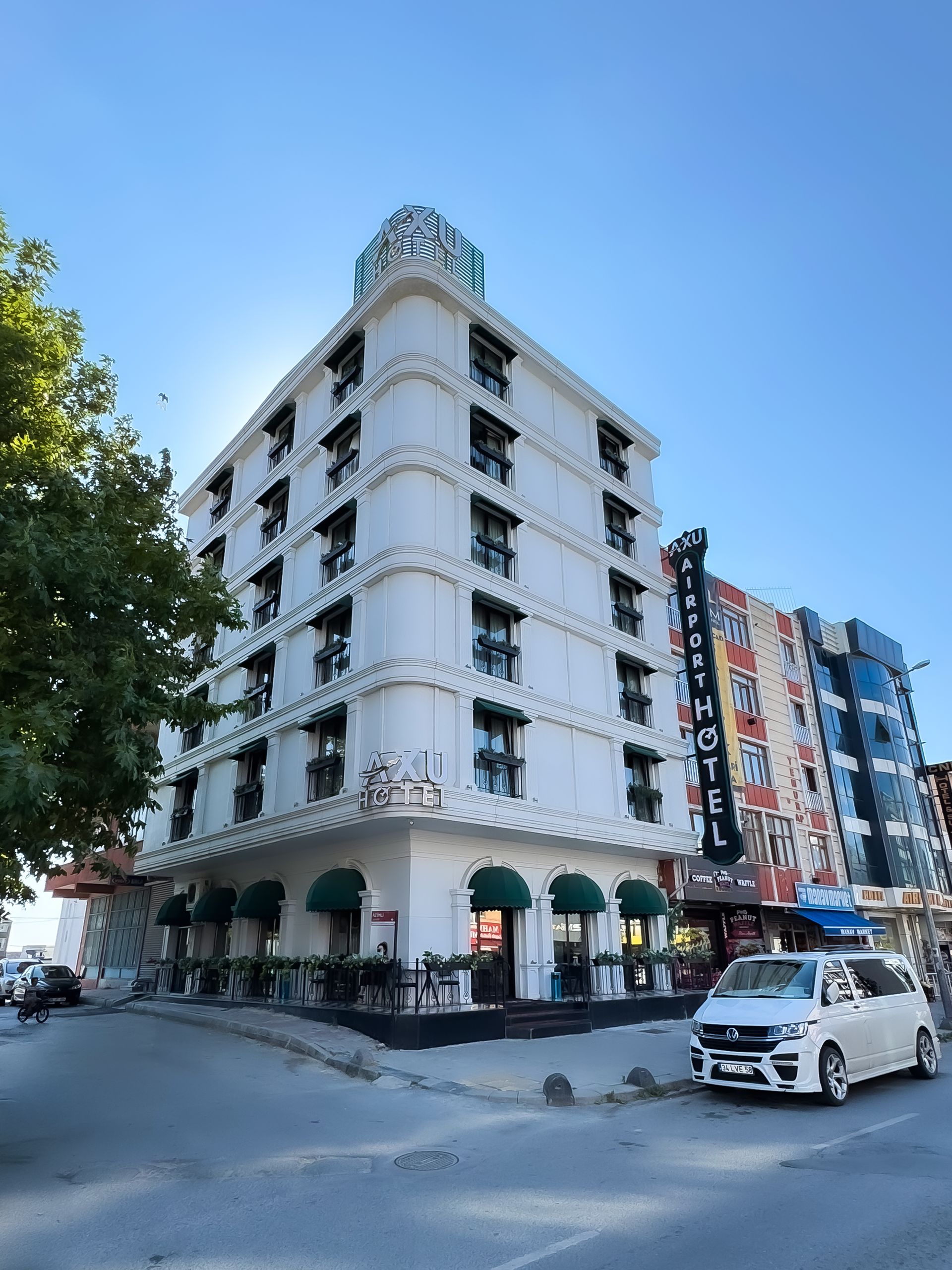 White corner building with green awnings on a sunny street, beside a blue hotel sign and parked white car.