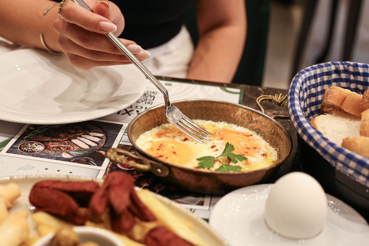Person stirring a skillet of eggs at a breakfast table with bread and coffee cups
