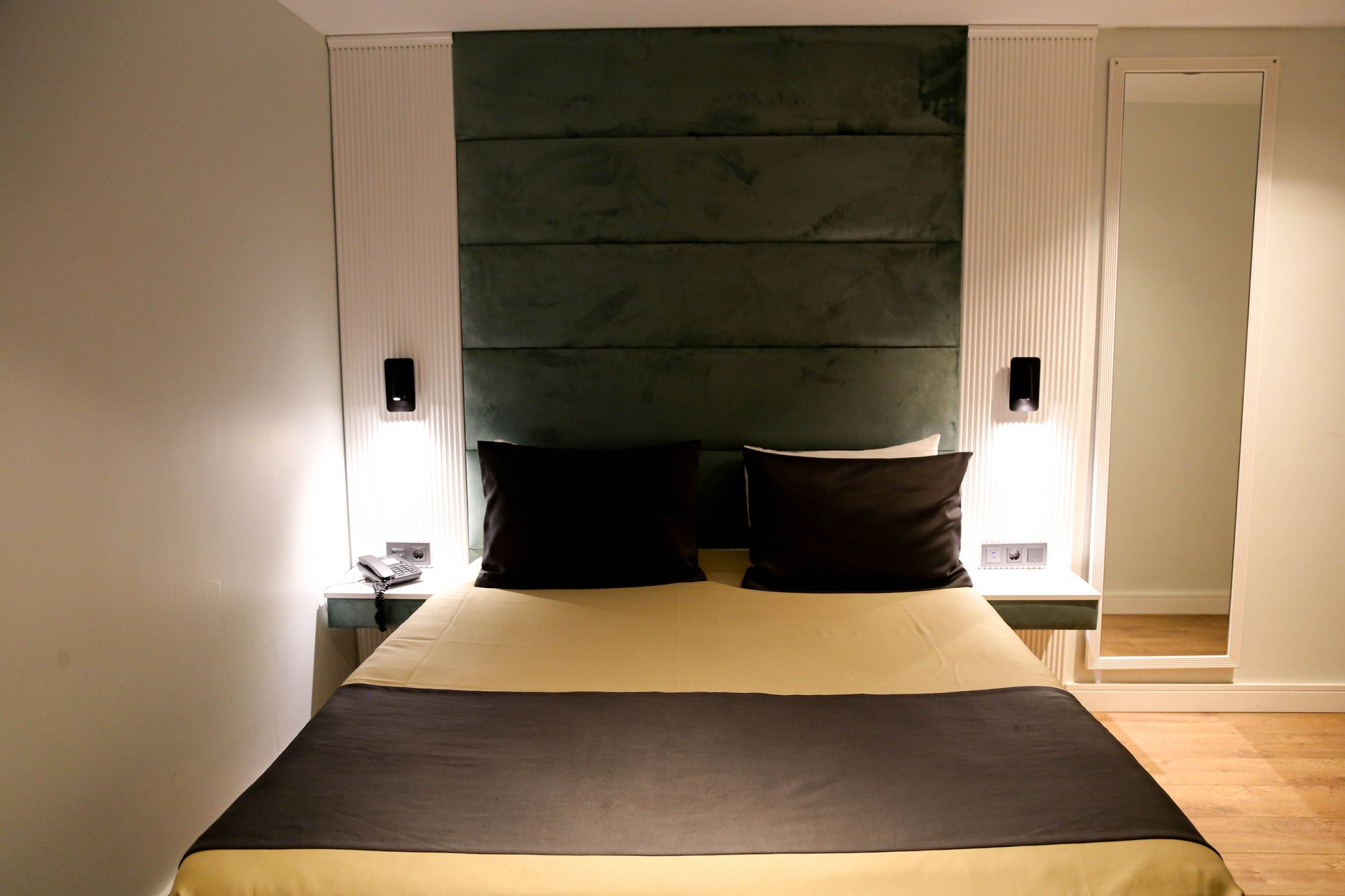Modern bedroom with a dark bed, black pillows, bedside lamps, and a green upholstered headboard panel