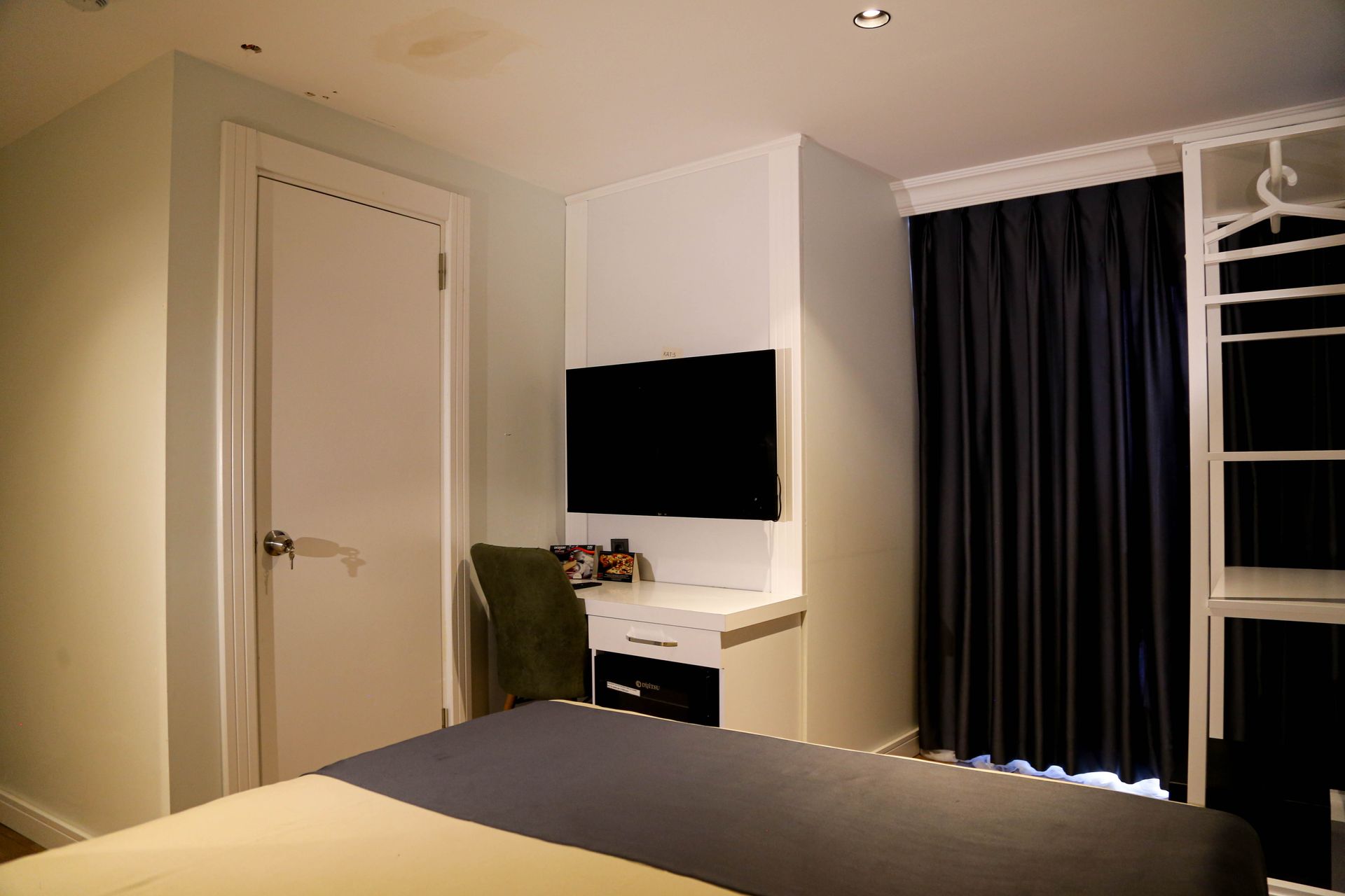 Modern hotel room with bed, wall-mounted TV, desk, chair, and black curtains by the window